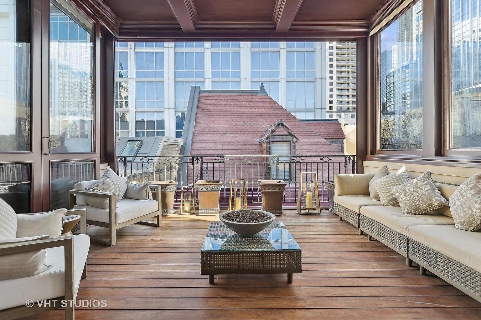 This image showcases a luxurious outdoor balcony or patio area, furnished with comfortable seating including a sofa and armchairs, arranged around a glass-topped coffee table. The space is enclosed with a railing and features potted plants and decorative lanterns, creating a cozy and inviting atmosphere. The backdrop reveals a cityscape with various buildings, suggesting an urban setting.