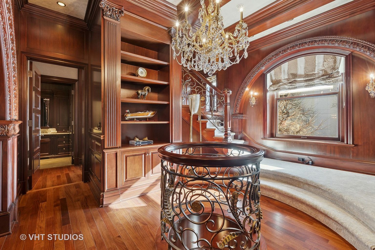 This interior shot showcases a luxurious hallway and staircase area, characterized by rich wood paneling and flooring. A prominent feature is the ornate wrought iron railing surrounding a circular opening, complemented by a grand crystal chandelier overhead. The space also includes built-in bookshelves and a window seat, creating an elegant and inviting atmosphere.