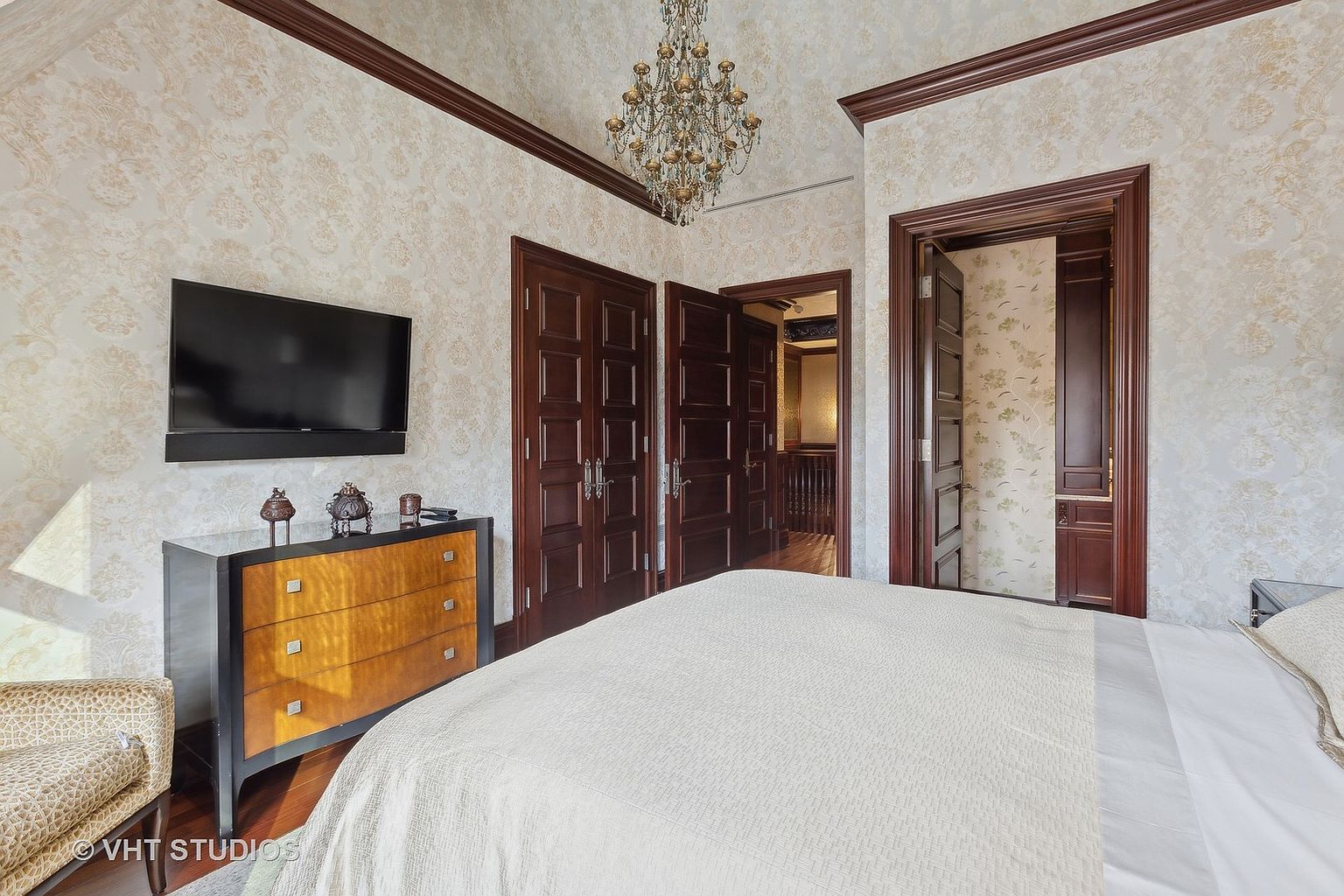 This is a primary bedroom featuring a bed with white linens, a dark wood dresser with a flat screen television mounted above it, and a decorative chandelier. The walls are covered in a patterned wallpaper, and the room has dark wood trim around the doors and ceiling. The overall style is traditional and luxurious.