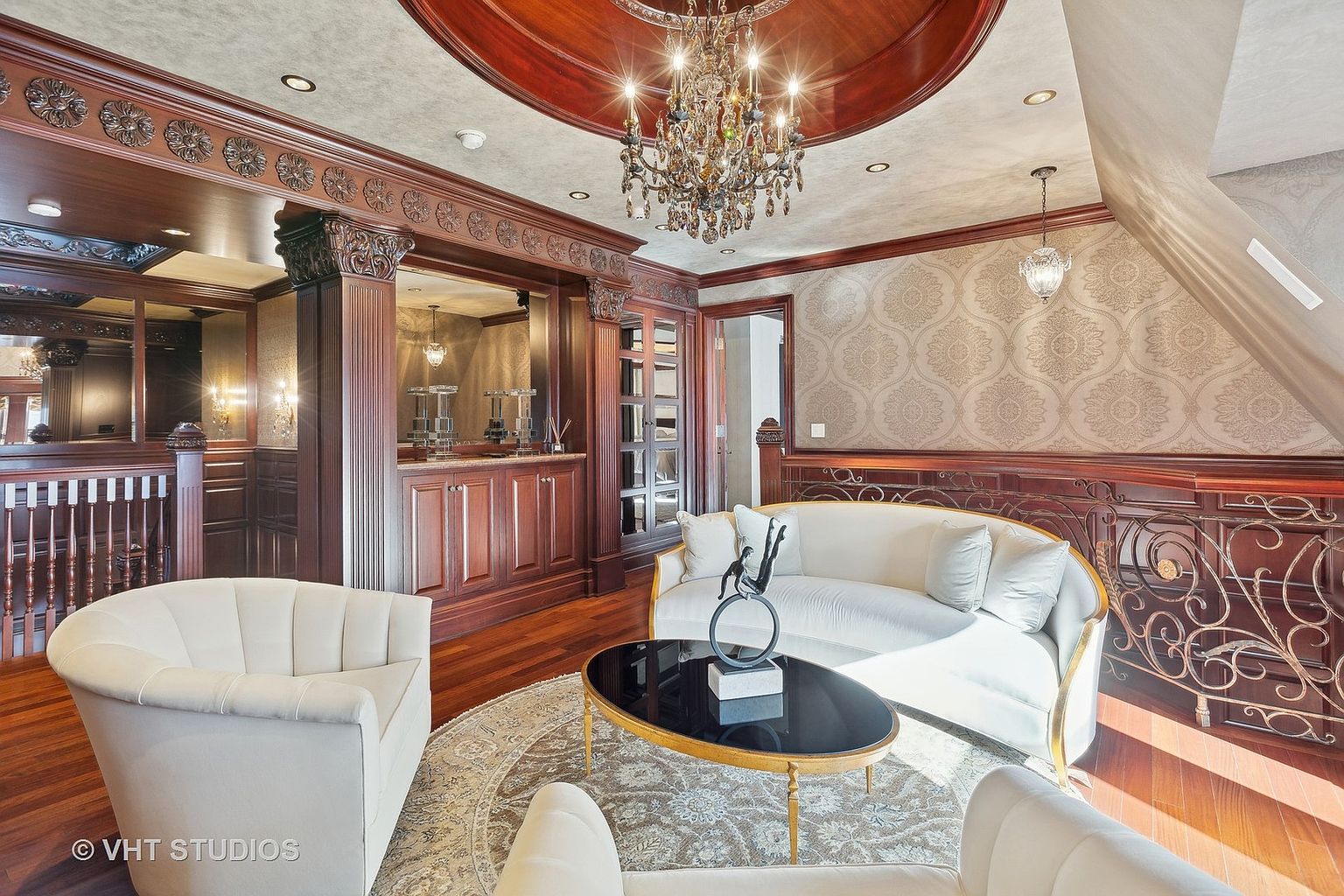 This is an elegant living room featuring rich wood detailing, including ornate trim and a coffered ceiling with a chandelier. The room is furnished with a white sofa, armchairs, and a black coffee table on a patterned rug. The space exudes a luxurious and sophisticated ambiance, perfect for formal entertaining or relaxation.