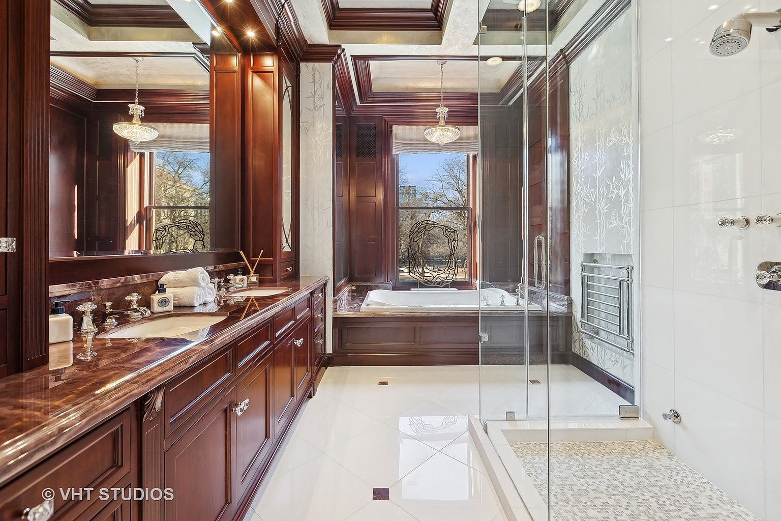 This is a luxurious primary bathroom featuring dark wood cabinetry with a marble countertop and dual sinks. A large window provides natural light above a soaking tub, and a glass-enclosed shower is visible to the right. The white tile flooring and walls contrast with the dark wood, creating a sophisticated and elegant space.