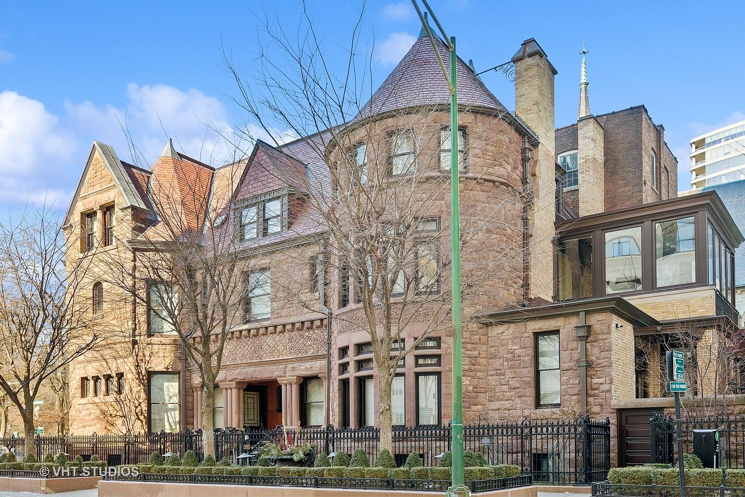 This is a front view of a grand, multi-story stone mansion with a turret and intricate architectural details. A wrought-iron fence surrounds the property, adding to its stately appearance. The building exudes a sense of timeless elegance and luxury, making it a highly desirable residence.