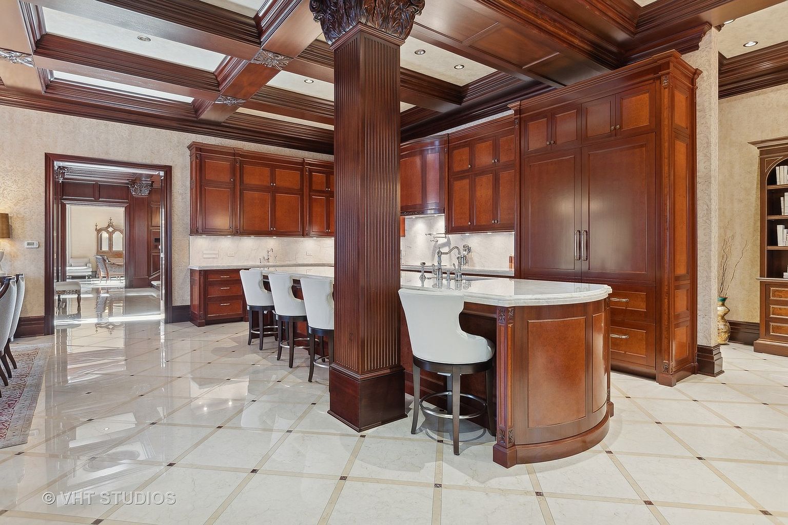 This is a luxurious kitchen featuring rich, dark wood cabinetry and a large central island with white marble countertops. The flooring is a light-colored tile with dark accents, and the ceiling has intricate wood beam detailing. The space is well-lit and exudes a sense of classic elegance and high-end craftsmanship.