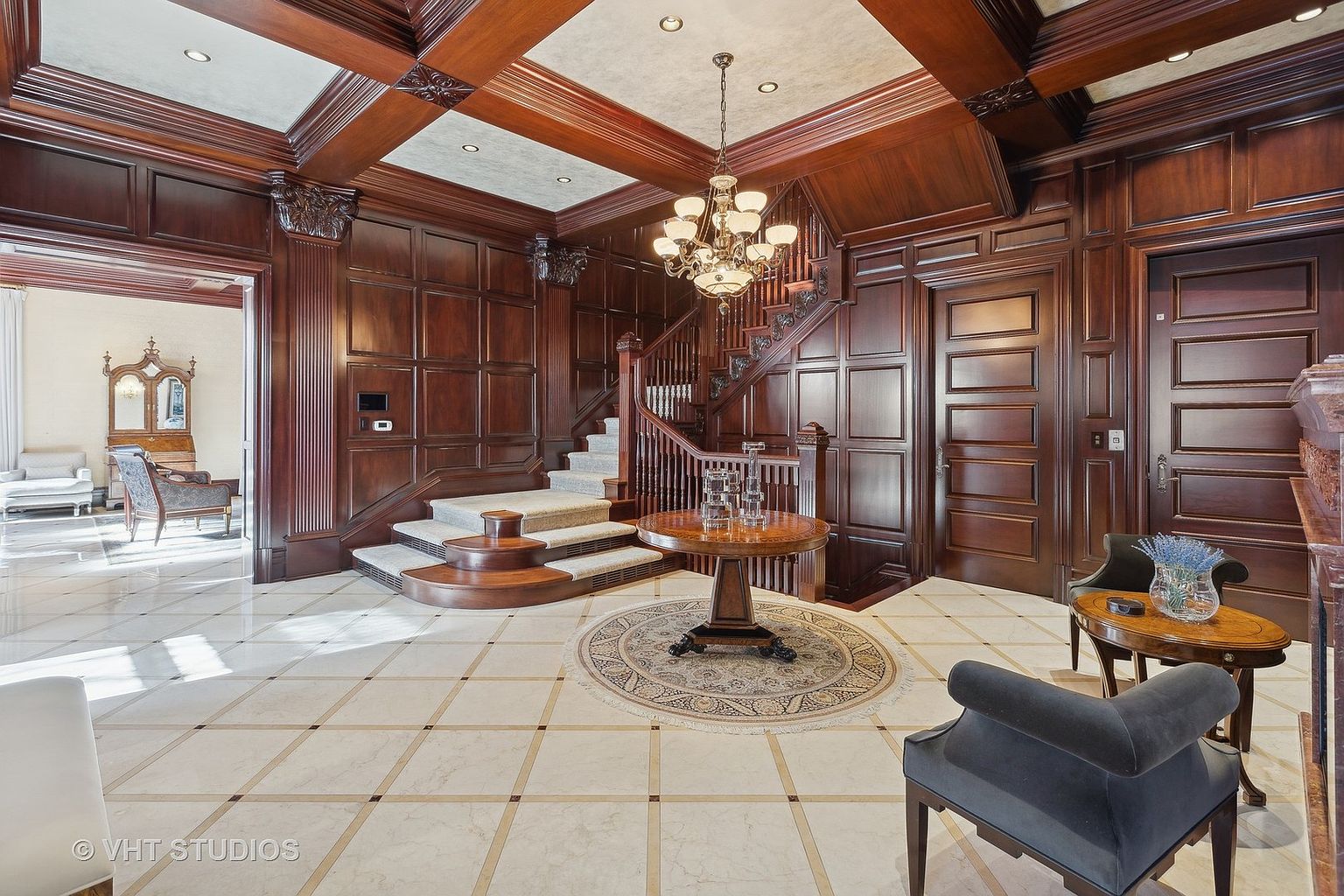 This grand foyer features rich wood paneling, a winding staircase with carpeted steps, and a striking chandelier. A round table with decorative glass objects sits atop a circular rug, adding to the luxurious feel. The marble tile flooring with accent inlays complements the overall elegant and traditional style of the home.
