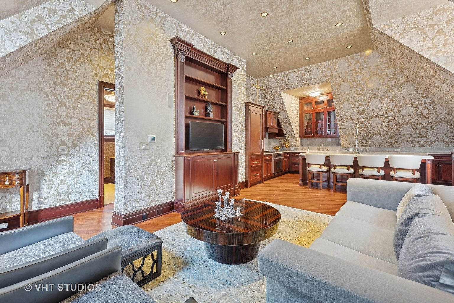 This interior shot showcases a luxurious living room with a partial kitchen area. The room features elegant wallpaper, rich wood flooring, and custom wood cabinetry with a built-in TV. A comfortable gray sofa and armchairs are arranged around a unique round coffee table, creating a sophisticated and inviting space.