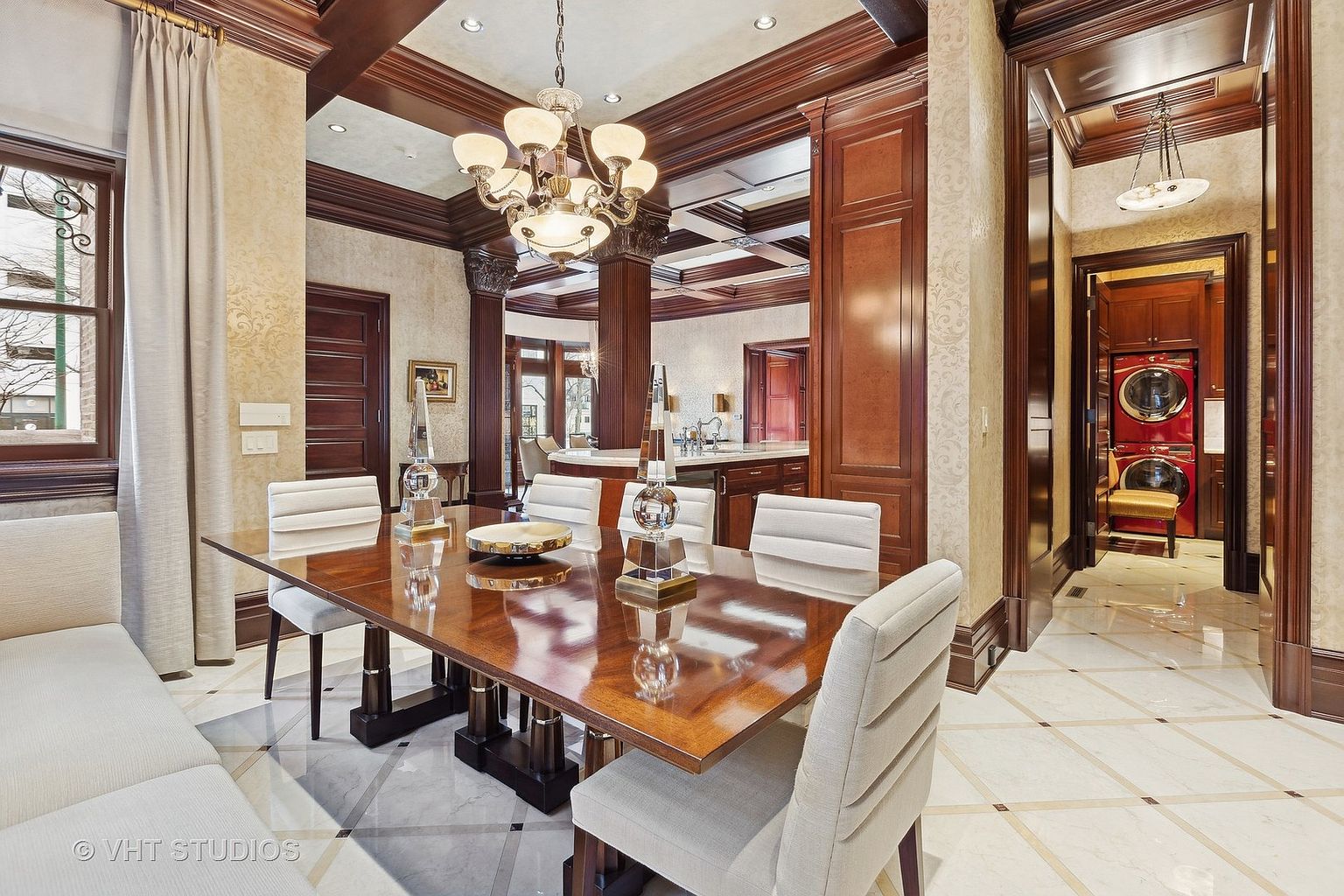 This interior shot showcases a luxurious dining room featuring a polished wood dining table with seating for six, complemented by elegant crystal centerpieces. The room is adorned with rich wood trim, patterned wallpaper, and a sophisticated chandelier, creating a warm and inviting atmosphere. A glimpse into the adjacent kitchen and laundry room adds depth and context to the home's layout.