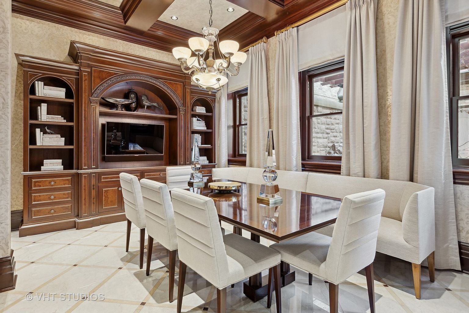 This is an interior shot of a formal dining room. The room features a large wooden table surrounded by upholstered chairs and a built-in bench. A chandelier hangs above the table, and a large wooden cabinet with shelving and a television is built into the wall. The flooring is tiled in a diamond pattern, and the walls are decorated with patterned wallpaper. The overall impression is one of luxury and sophistication.
