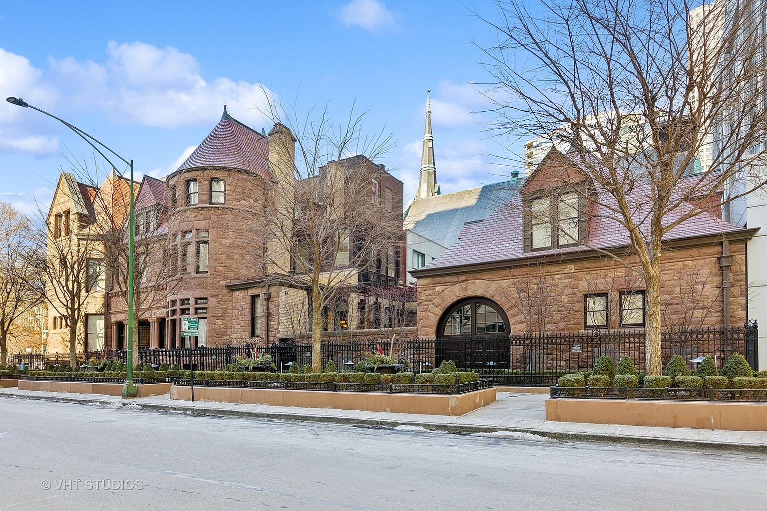This image showcases the front exterior of a grand, historic building constructed of brown stone. The architecture features intricate detailing, including a turret, arched windows, and a wrought-iron fence enclosing well-manicured landscaping. The scene is set on a clear day, highlighting the building's impressive facade and street presence.