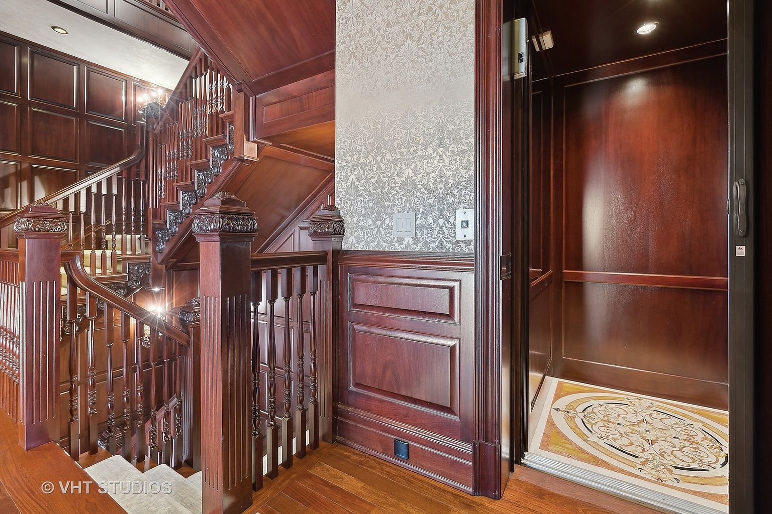 This interior shot showcases a grand wooden staircase with intricate detailing and a classic design. Adjacent to the staircase is an open elevator, revealing a decorative floor. The rich wood tones and elegant architectural features create a luxurious and sophisticated atmosphere.
