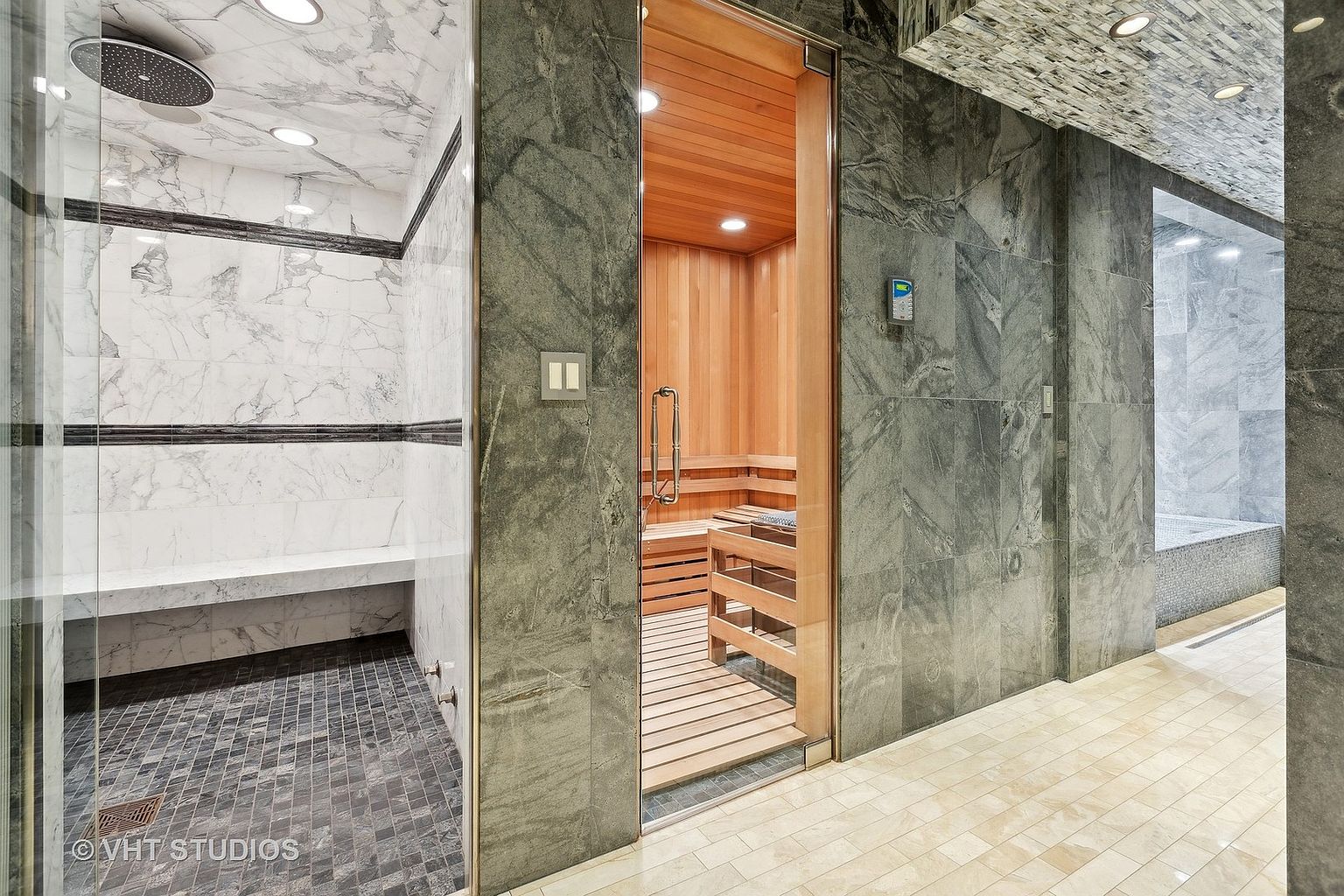 This image showcases a luxurious primary bathroom featuring a walk-in shower with marble tiling and a built-in bench, a sauna with wooden interiors, and a separate bathing area. The design incorporates a sophisticated color palette of gray and white, creating a spa-like atmosphere. The perspective is from the doorway, highlighting the seamless integration of these amenities.