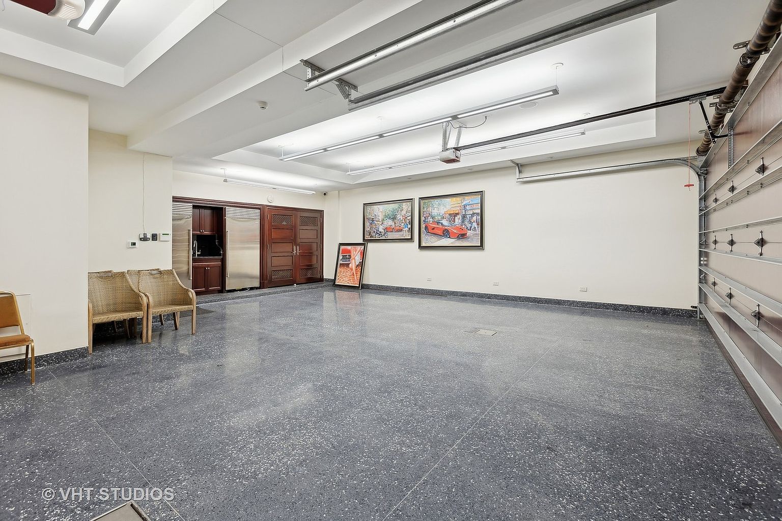 This is a spacious and well-lit garage featuring a polished, speckled floor and a high ceiling with industrial lighting fixtures. The garage includes built-in cabinetry and a refrigerator, suggesting potential for use as a multi-purpose space. Artwork adorns the walls, adding a touch of sophistication to the functional area.