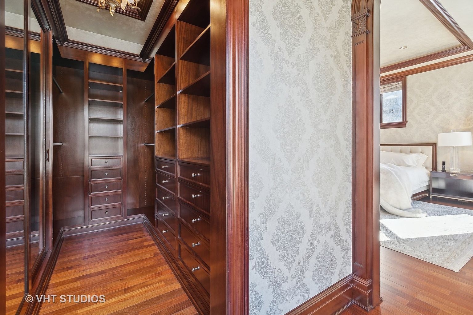 This image showcases a luxurious walk-in closet with rich, dark wood cabinetry and shelving. The closet features ample storage space with drawers and shelves, and the hardwood flooring adds warmth. Through the doorway, a glimpse of a bedroom with elegant wallpaper and a bed can be seen, suggesting a seamless transition between spaces.
