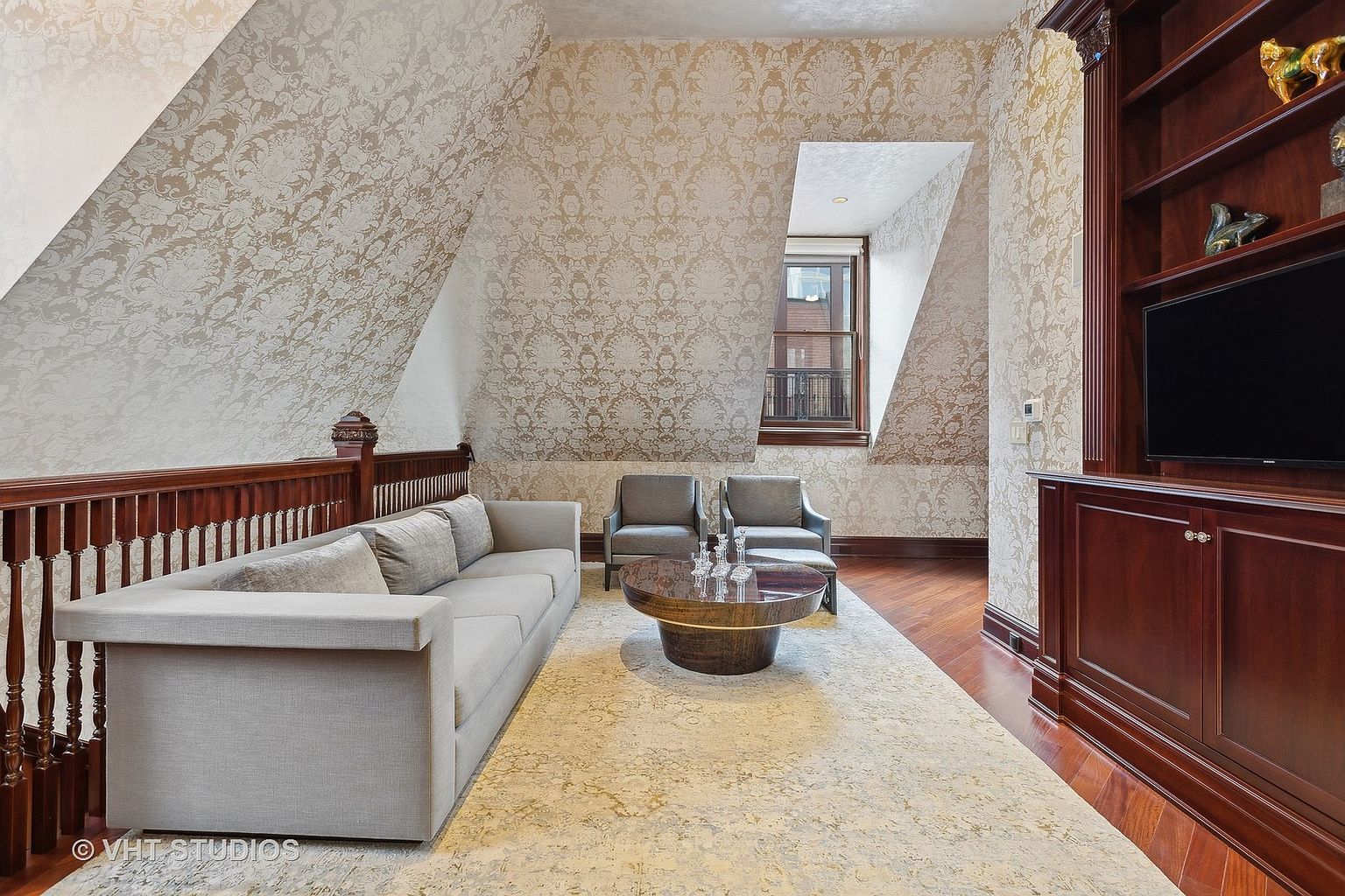 This is an interior shot of a living room featuring a long gray sofa, two gray armchairs, and a round coffee table on a patterned rug. The walls are covered in a patterned wallpaper, and there is a built-in wooden cabinet with a television. The room has an attic-like feel with angled walls and a small window.