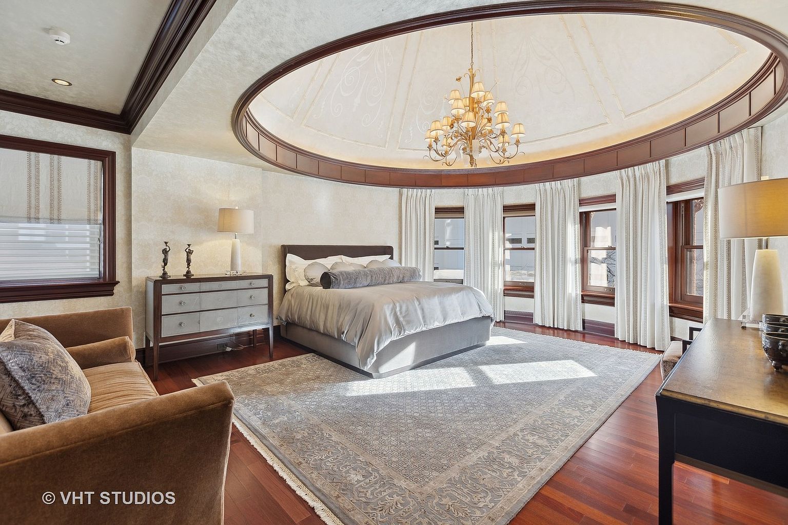This is a luxurious primary bedroom featuring a unique circular ceiling with a decorative chandelier. The room is well-lit with natural light streaming through multiple windows framed by elegant white curtains. A large area rug covers the hardwood floors, and the room is furnished with a bed, dresser, and seating area, creating a comfortable and sophisticated space.