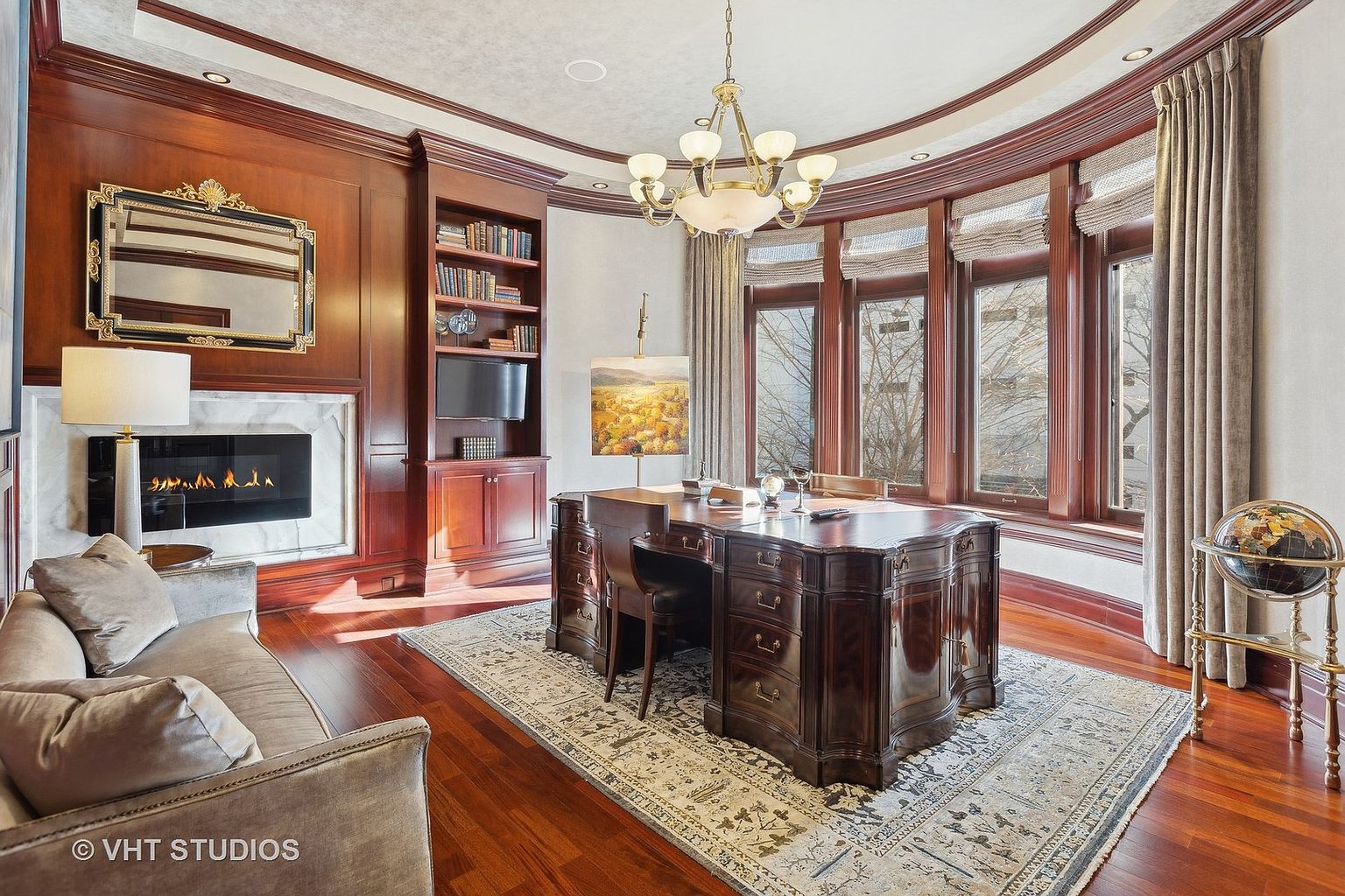 This is a luxurious home office featuring rich wood paneling, a large executive desk, and a cozy fireplace. The room is well-lit by natural light from a bay window and an elegant chandelier. A comfortable sofa and a decorative globe add to the room's sophisticated and inviting atmosphere.