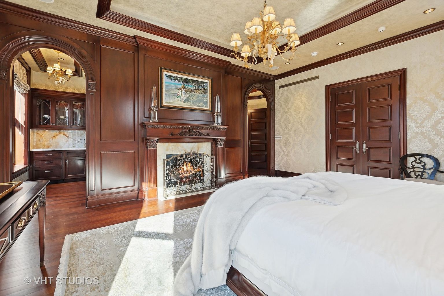 This is a luxurious primary bedroom featuring rich wood paneling, a fireplace with an ornate screen, and a grand chandelier. The room includes a doorway leading to another room and a double door, suggesting ample space and privacy. A large bed with white linens dominates the foreground, adding to the room's opulent and inviting atmosphere.