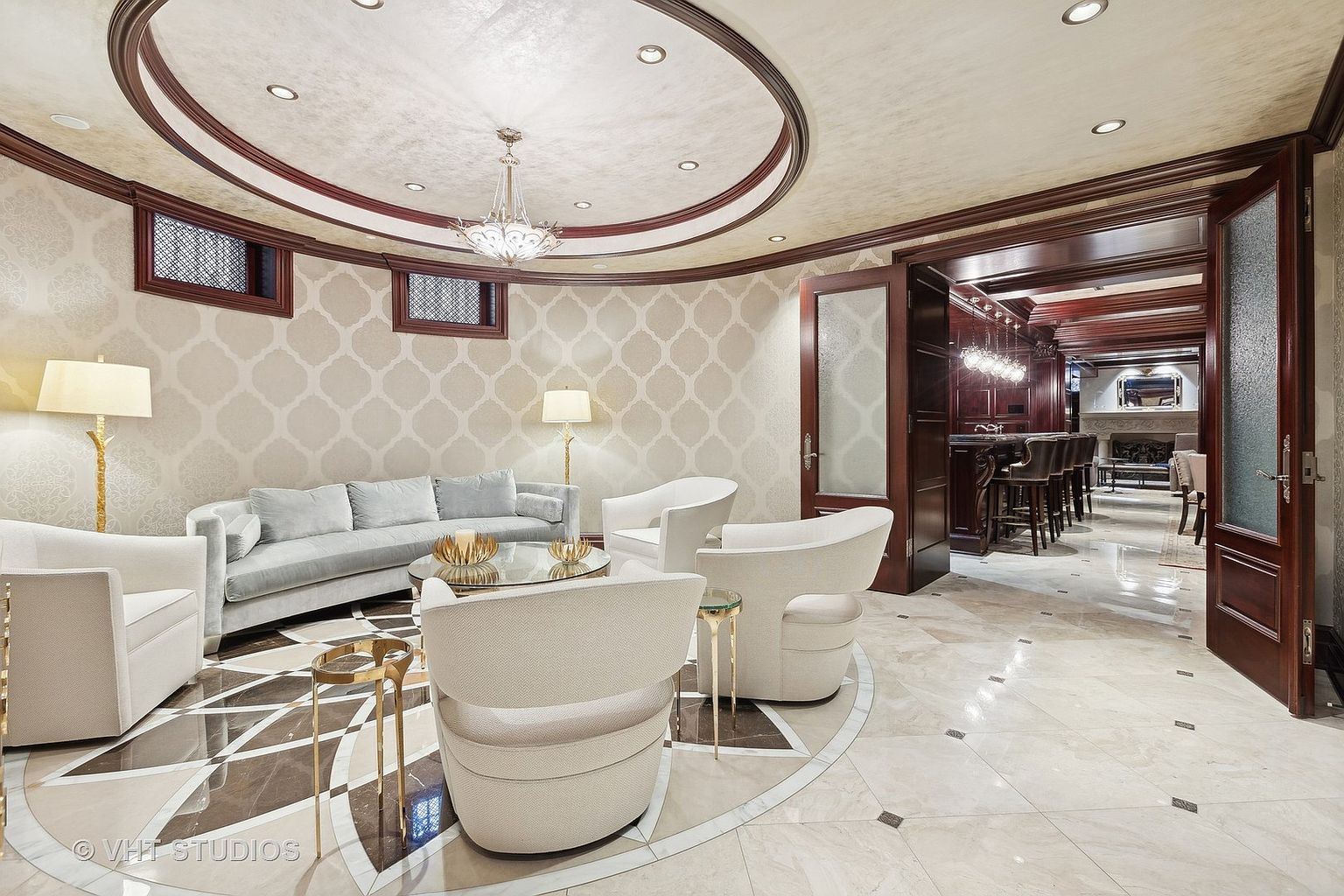 This is an interior shot of a luxurious living room featuring a curved wall with patterned wallpaper, a light gray sofa, and several white armchairs arranged around a glass coffee table. The room boasts a unique ceiling design with a circular recessed area and a chandelier, complemented by a striking geometric tile floor. An open doorway leads to a bar area, enhancing the room's entertainment potential.