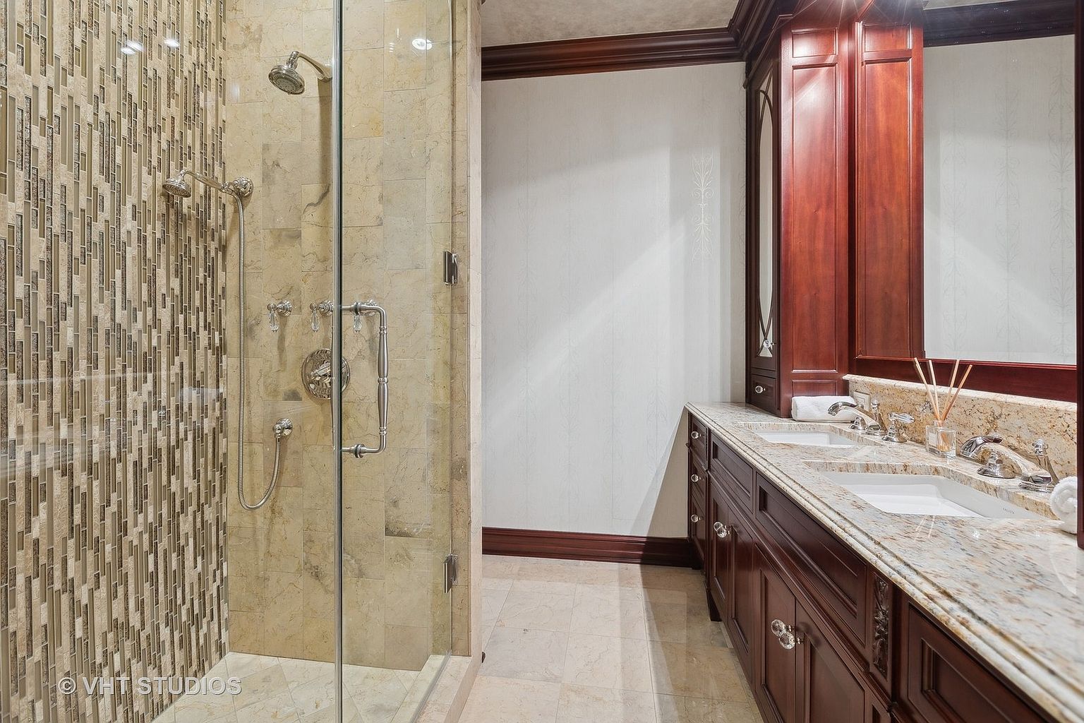 This is a primary bathroom featuring a glass-enclosed shower with multiple shower heads and a dual vanity with a granite countertop. The vanity has dark wood cabinetry with decorative hardware. The walls are a light color with subtle patterns, and the flooring is tiled. The perspective is from the doorway, showcasing the shower on the left and the vanity on the right.
