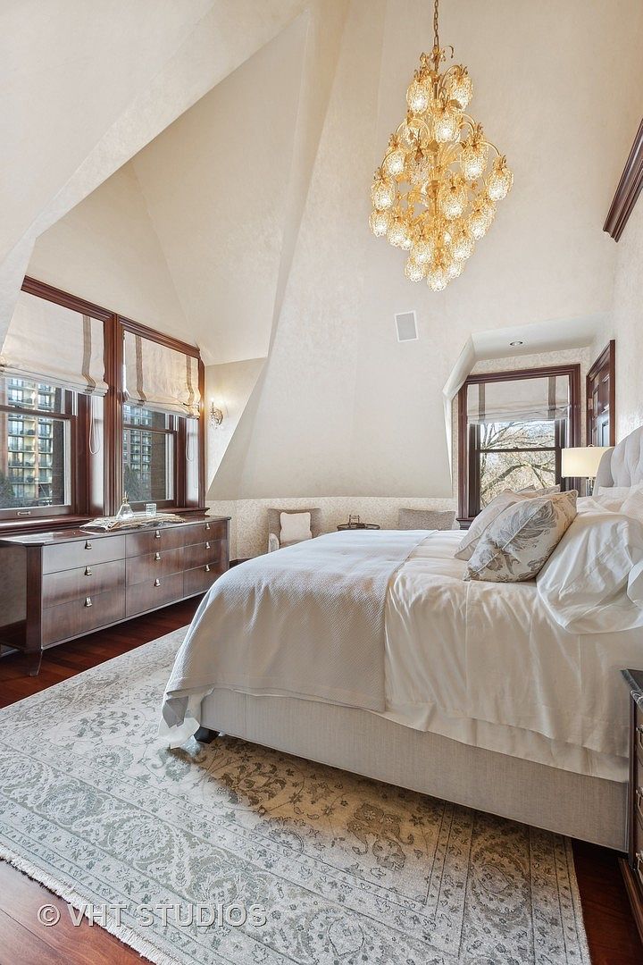 This is a primary bedroom featuring a large bed with white linens, a decorative rug, and a dresser with windows above. The room has an elegant chandelier and an angled ceiling, creating a unique architectural detail. The overall impression is luxurious and comfortable.