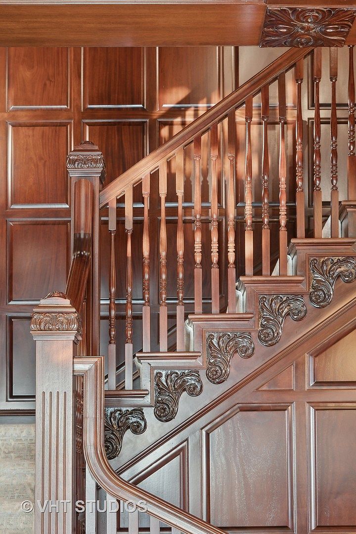 This image showcases a beautifully crafted wooden staircase with intricate carvings and paneling. The rich, dark wood creates a sense of luxury and timeless elegance. The staircase is well-lit, highlighting the detailed craftsmanship and adding warmth to the space, making it a focal point of the home.
