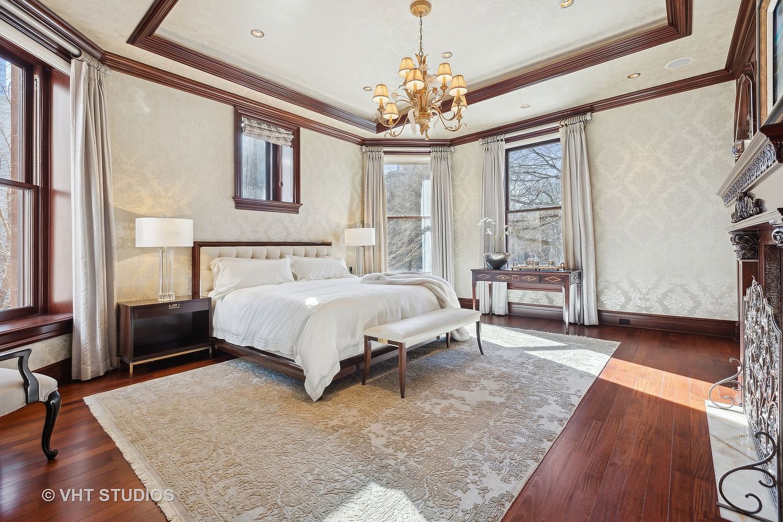 This is a luxurious primary bedroom featuring a large bed with white linens, flanked by dark wood nightstands and lamps. The room is decorated with elegant wallpaper, dark wood trim, and a chandelier, creating a sophisticated and comfortable atmosphere. A large area rug covers most of the hardwood floor, and natural light streams in through the windows.