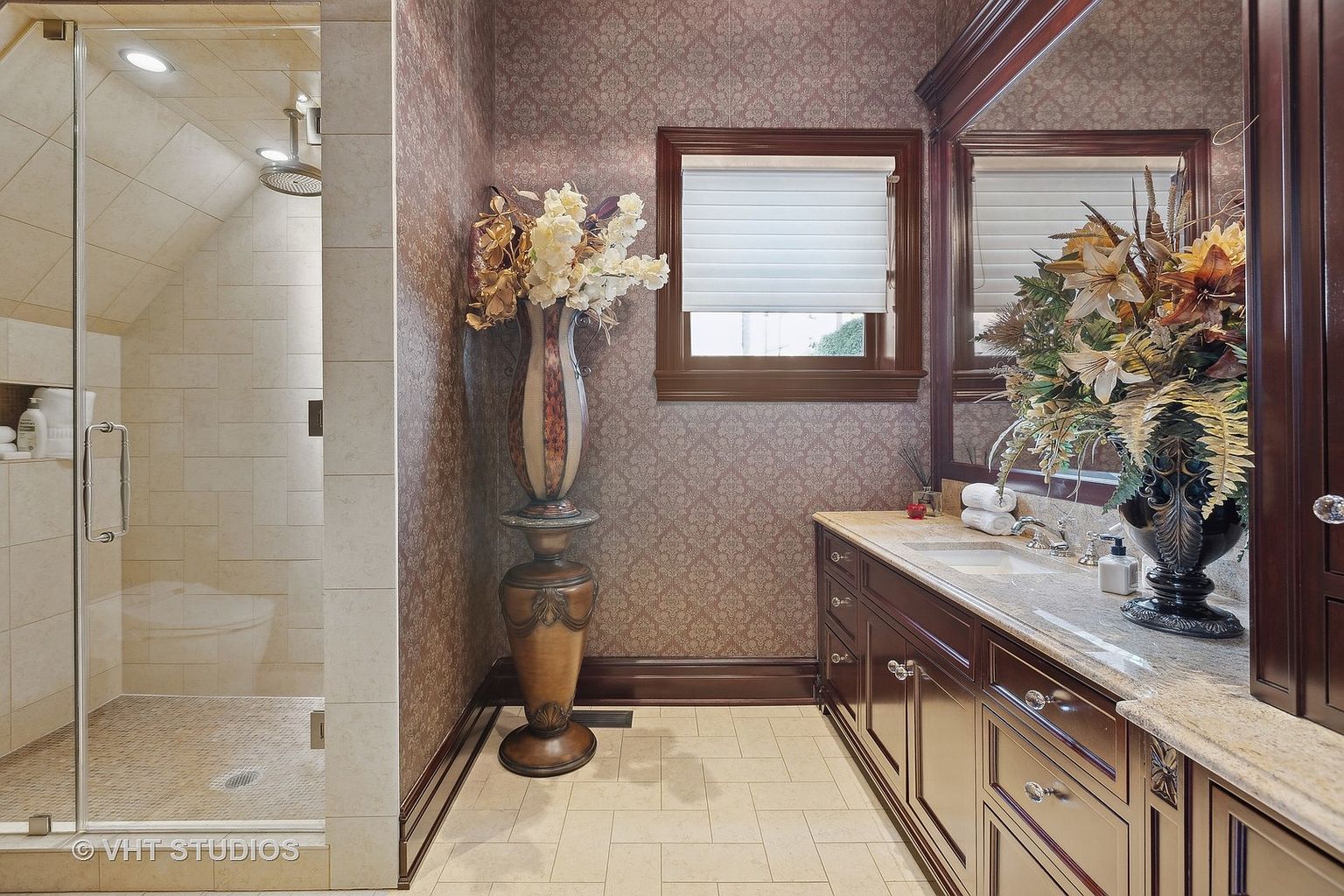 This is a well-appointed primary bathroom featuring a glass-enclosed shower with beige tiling, a dark wood vanity with a light-colored countertop, and decorative floral arrangements. The walls are covered in patterned wallpaper, and the flooring is light tile. The overall impression is luxurious and elegant.