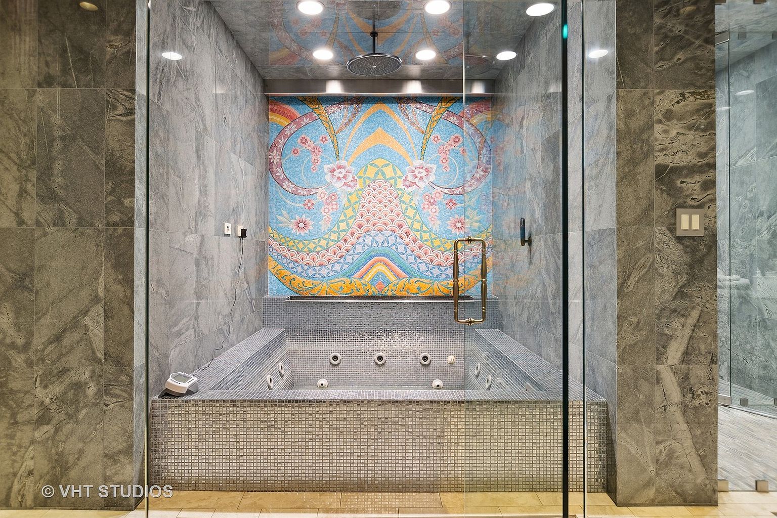 This is a luxurious primary bathroom featuring a mosaic-tiled jacuzzi tub enclosed by glass walls. The walls are covered in gray marble, and the ceiling has recessed lighting. A colorful mosaic mural adorns the wall behind the tub, adding an artistic focal point to the space.
