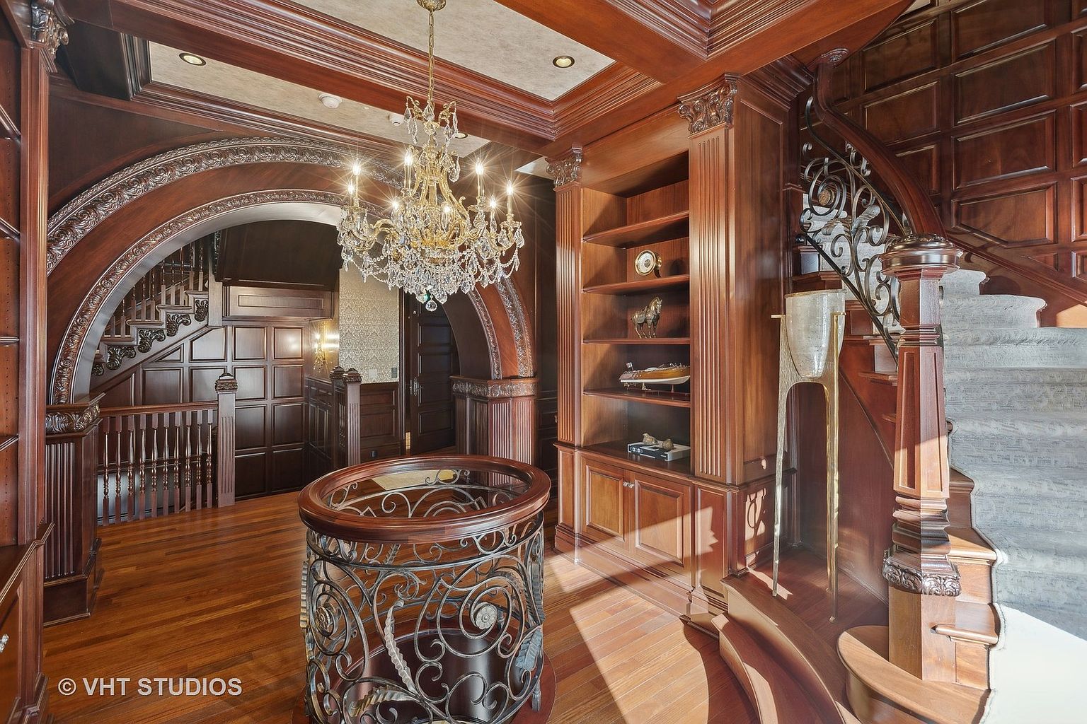 This grand interior hallway features rich wood paneling, a stunning crystal chandelier, and an ornate wrought iron railing. A curved staircase leads to the upper level, while a built-in bookshelf adds character and functionality. The space exudes luxury and timeless elegance, perfect for a high-end real estate listing.