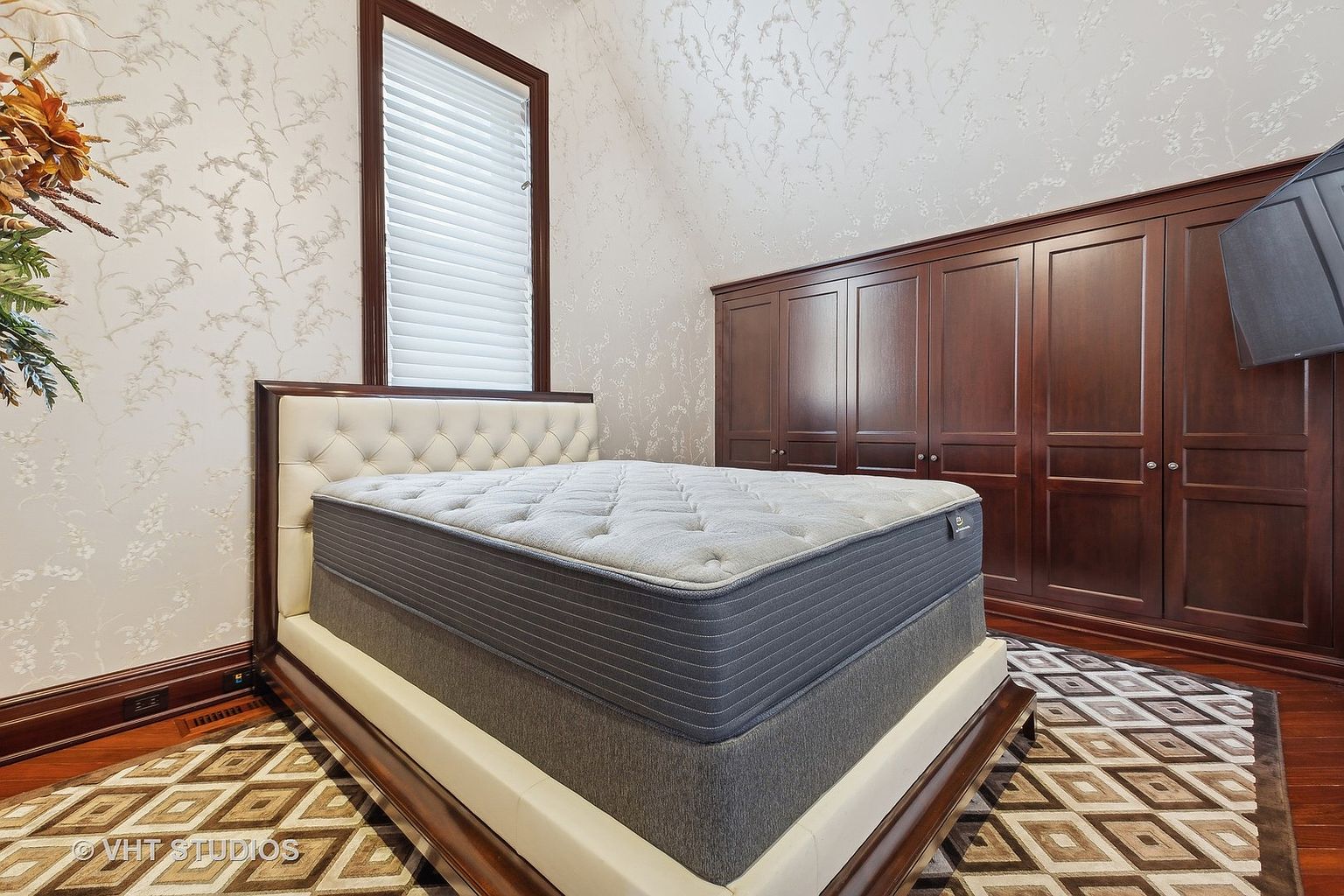 This is a well-appointed primary bedroom featuring a large bed with a tufted headboard and a plush mattress. The room includes a large, dark wood wardrobe and a patterned rug that adds visual interest. The walls are adorned with a subtle floral wallpaper, and natural light filters through a window with blinds, creating a serene and inviting atmosphere.