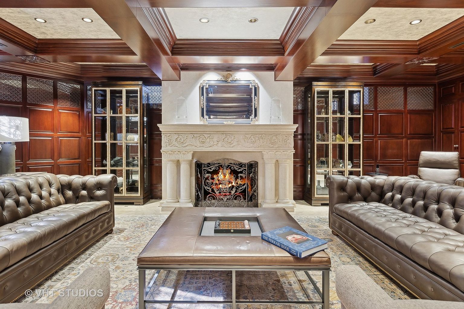 This is a luxurious living room featuring two brown leather Chesterfield sofas facing each other, flanking a large stone fireplace with an ornate metal screen. A leather ottoman with a metal frame sits in the center, adorned with books. The room is richly decorated with dark wood paneling, coffered ceilings, and glass-fronted display cabinets, creating a sophisticated and inviting atmosphere.
