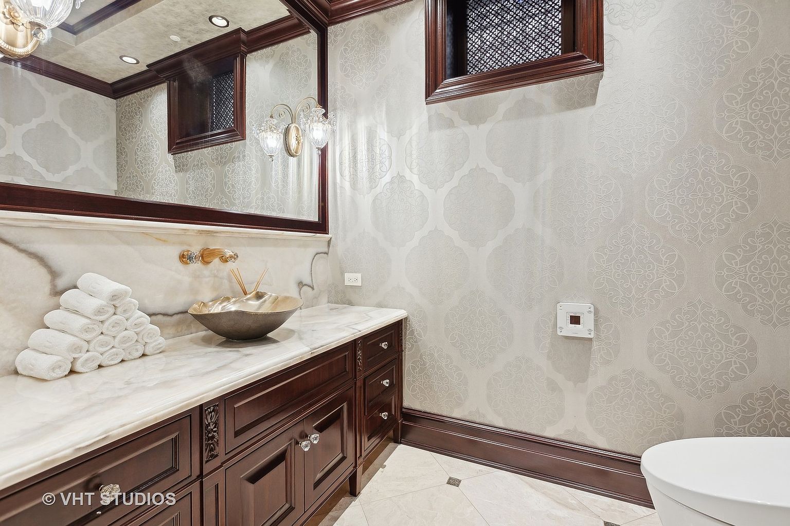 This is a well-appointed guest bathroom featuring a dark wood vanity with a marble countertop and a decorative silver bowl. The walls are adorned with patterned wallpaper, and a framed mirror hangs above the sink. The room exudes a sense of luxury and sophistication, making it an appealing feature for potential buyers.