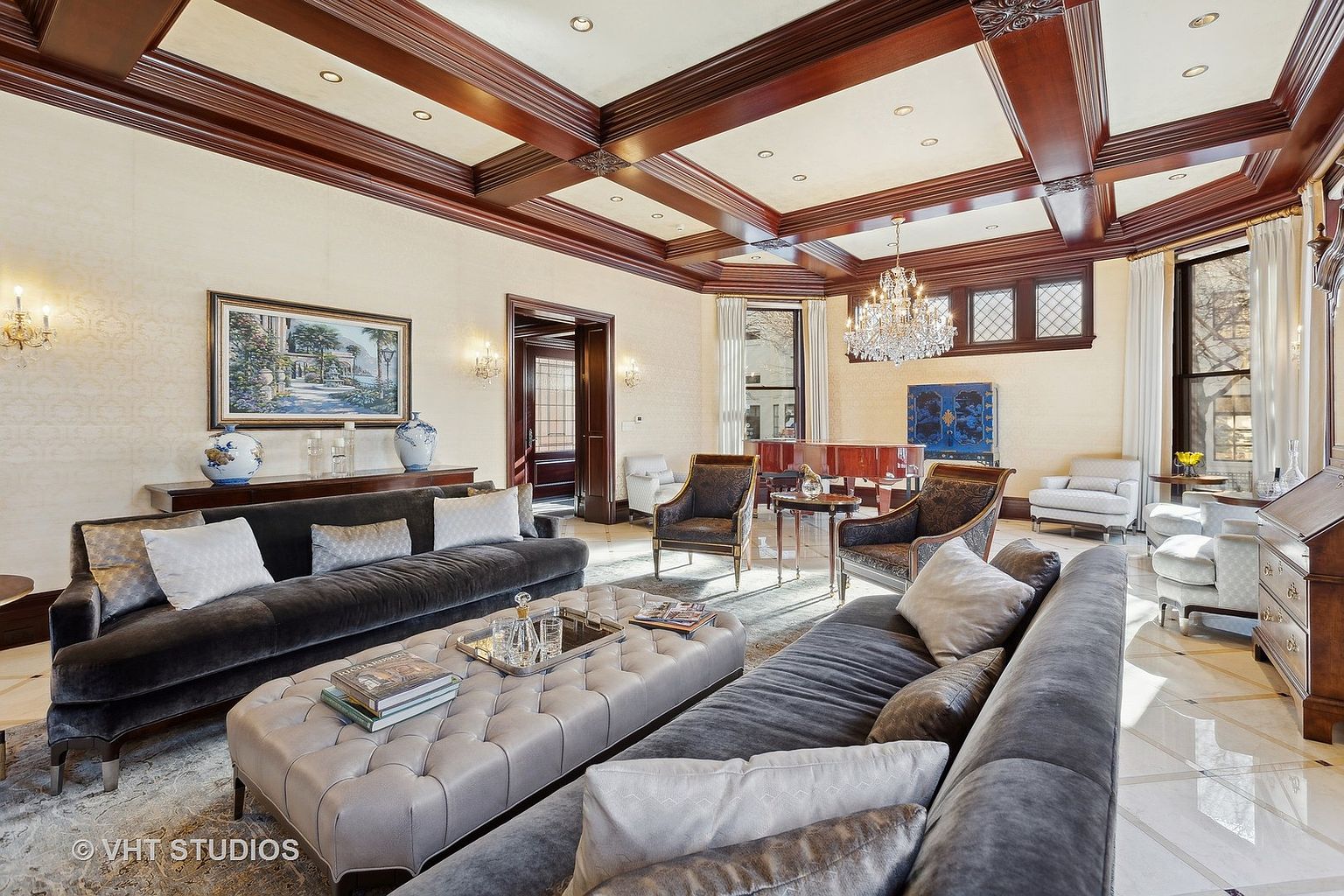 This is an interior shot of a luxurious living room featuring two plush gray sofas, a tufted ottoman, and elegant decor. The room is illuminated by a crystal chandelier and wall sconces, highlighting the detailed ceiling beams and the artwork on the walls. The overall impression is one of sophistication and comfort, perfect for a high-end real estate listing.