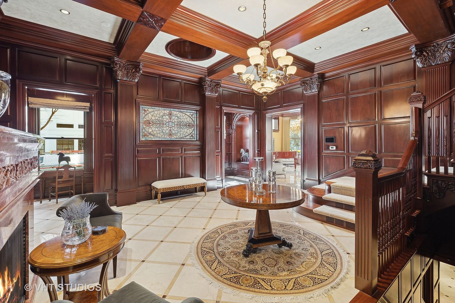 This grand foyer features rich, dark wood paneling and trim, creating a luxurious and traditional aesthetic. A circular table with decorative objects sits atop an ornate rug, while a staircase with detailed woodwork adds to the elegance. The space is illuminated by a multi-tiered chandelier, enhancing the opulent feel.
