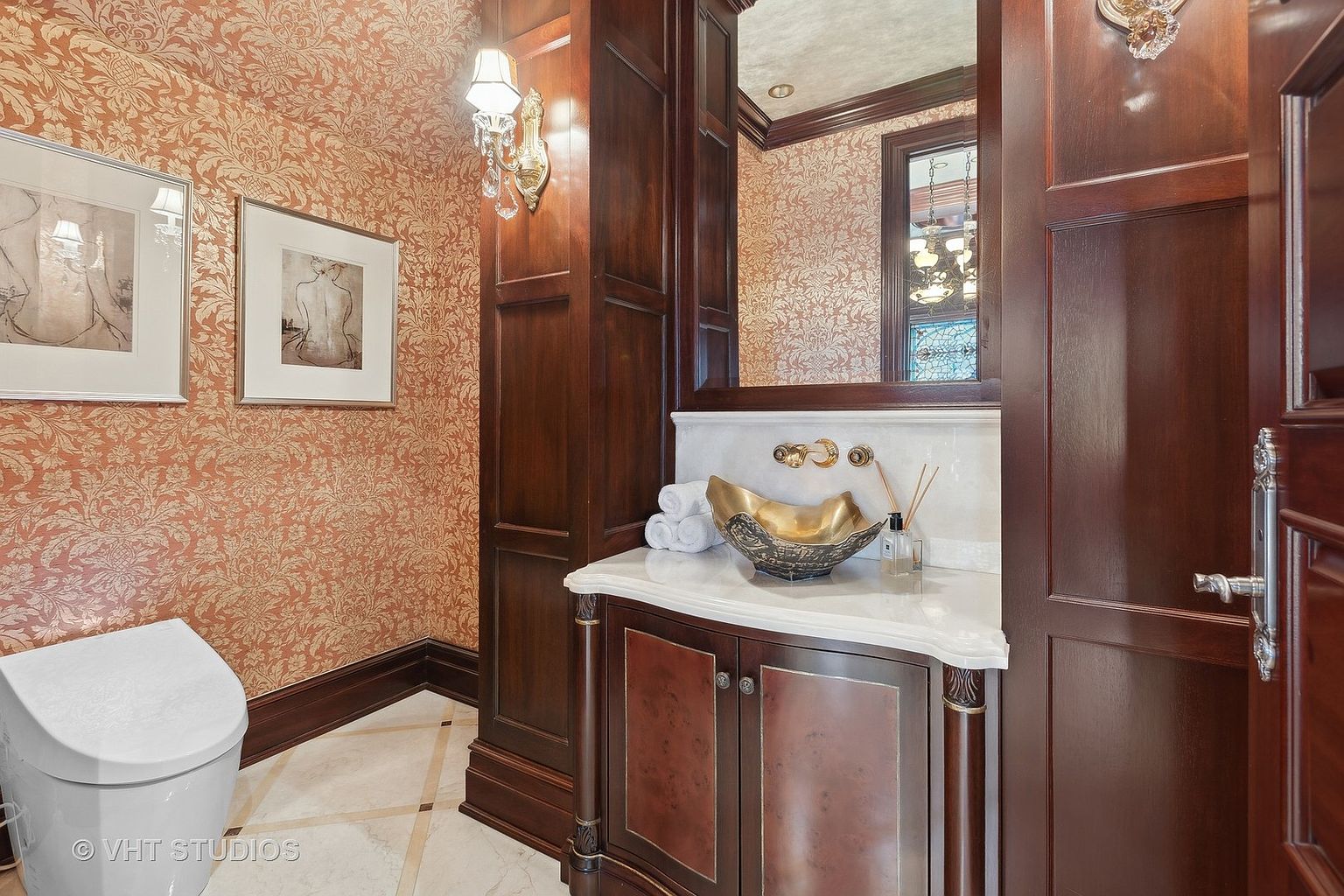 This is a well-appointed guest bathroom featuring a unique gold-toned vessel sink, marble countertops, and dark wood cabinetry. The walls are adorned with patterned wallpaper and framed artwork, adding a touch of elegance. The flooring is a light-colored tile with dark accents, and a partially visible toilet is present.