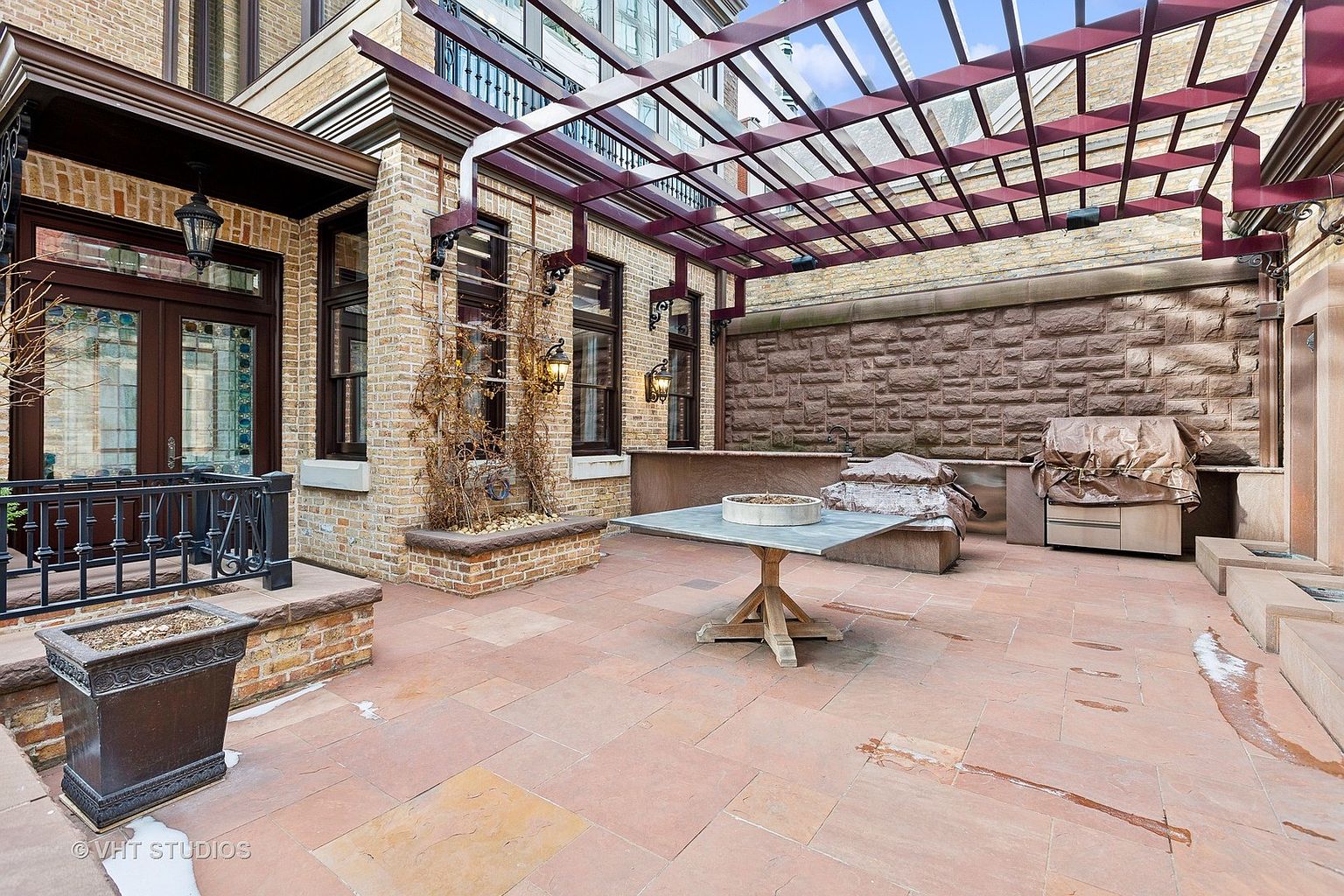 This is a beautifully designed outdoor patio area, featuring a brick-paved surface and a pergola overhead. The space includes a stone wall backdrop, a table, and a built-in grill area covered with a tarp. The overall impression is one of a luxurious and inviting outdoor living space.