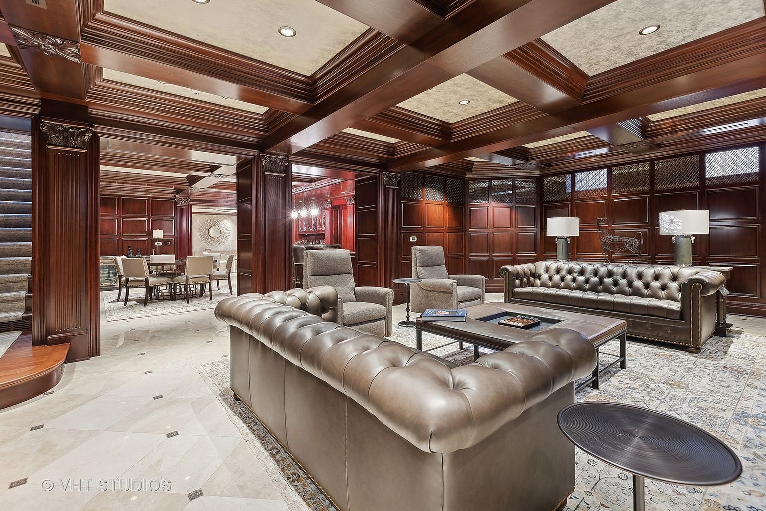 This is an interior shot of a luxurious living room featuring rich, dark wood paneling and coffered ceilings. The room is furnished with plush leather Chesterfield sofas and armchairs, arranged around a dark wood coffee table on an ornate rug. The space exudes a sophisticated and classic ambiance, ideal for entertaining or relaxing.