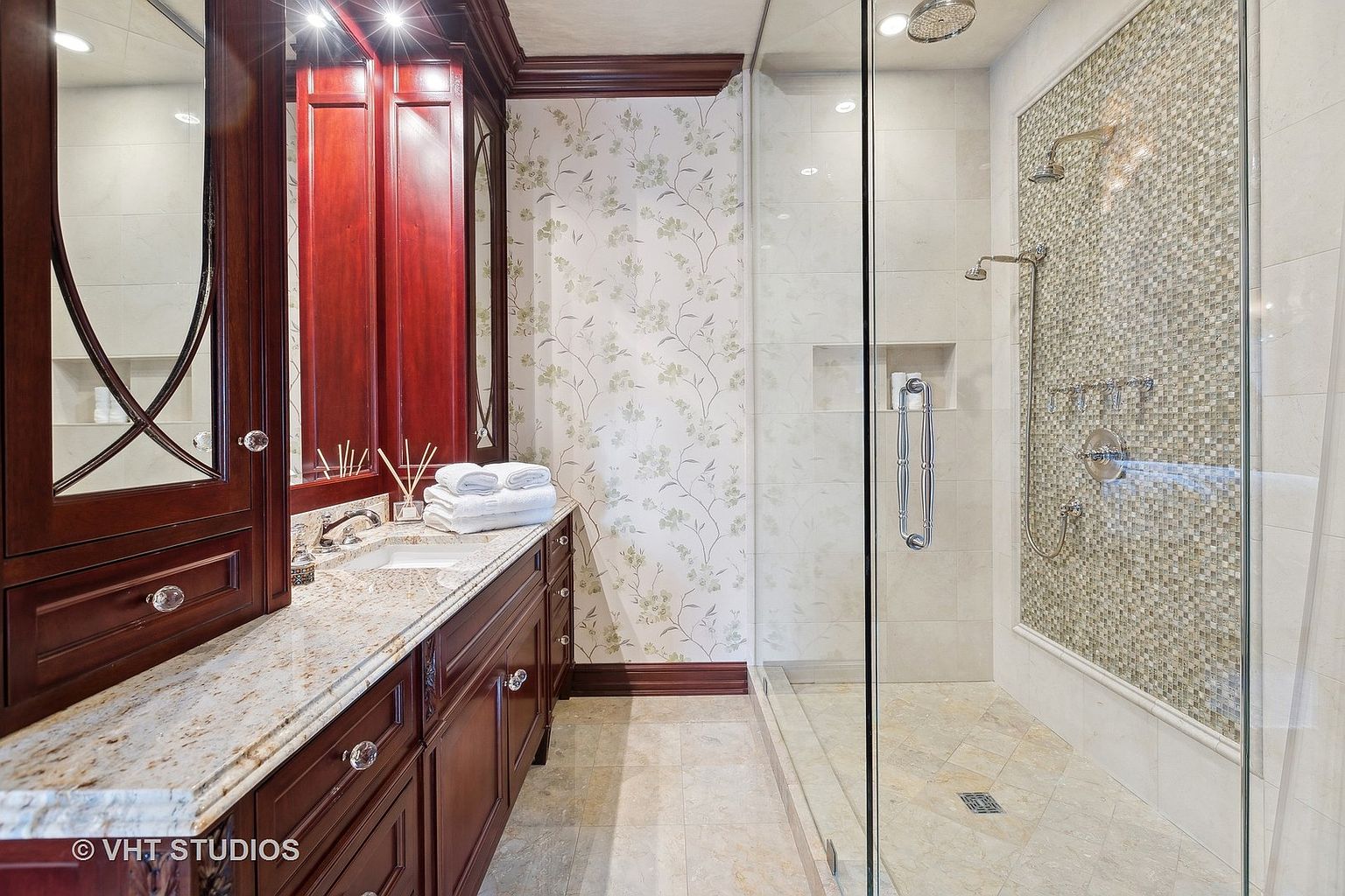 This is a well-appointed primary bathroom featuring a dark wood vanity with a light-colored granite countertop and crystal knobs. The walls are adorned with floral wallpaper, and a glass-enclosed shower showcases mosaic tile accents. The overall impression is luxurious and elegant.