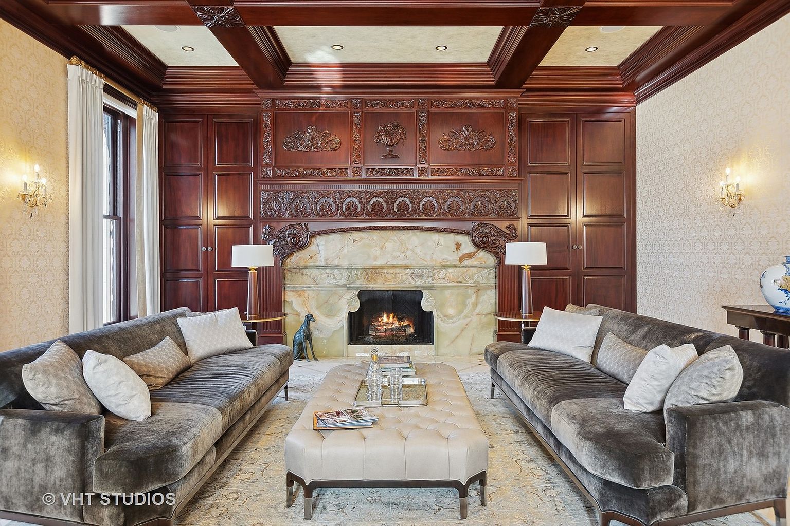 This is a luxurious living room featuring two plush gray sofas facing each other, flanking a tufted ottoman coffee table. A grand fireplace with intricate wood detailing and marble surround serves as the focal point, complemented by matching lamps on either side. The room exudes a sense of warmth and sophistication with its rich wood paneling and elegant decor.