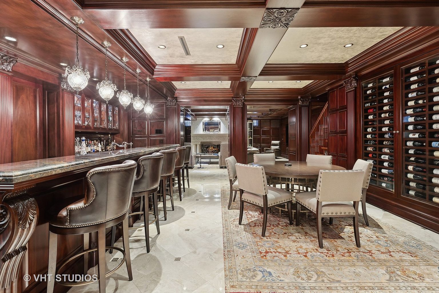 This is a luxurious basement featuring a bar area with seating, a dining area with a round table and chairs set on an ornate rug, and a wine cellar. The space is richly decorated with dark wood paneling, coffered ceilings, and elegant lighting fixtures, creating a sophisticated and inviting atmosphere. A fireplace is visible in the background, adding to the ambiance.