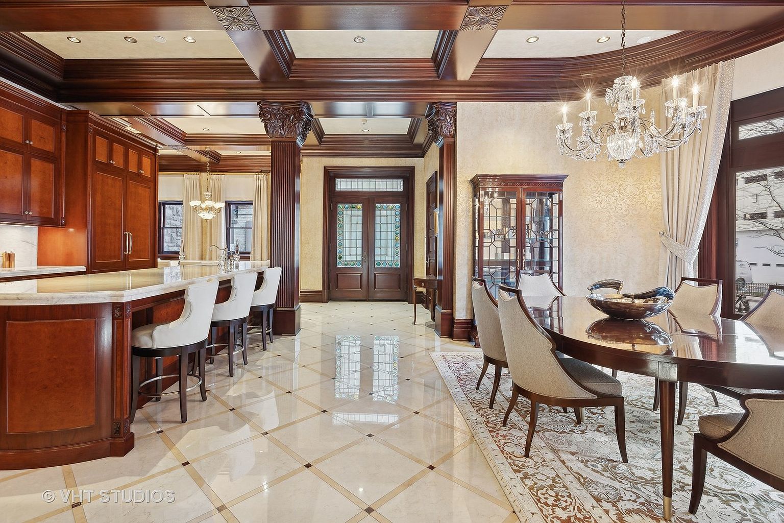 This interior shot showcases a luxurious dining area adjacent to a kitchen space, featuring rich wood finishes on the cabinetry, ceiling beams, and decorative columns. A grand chandelier hangs above a polished wood dining table surrounded by upholstered chairs, while a display cabinet adds to the room's elegance. The glossy tiled floor reflects light, enhancing the spacious and opulent feel of the room.