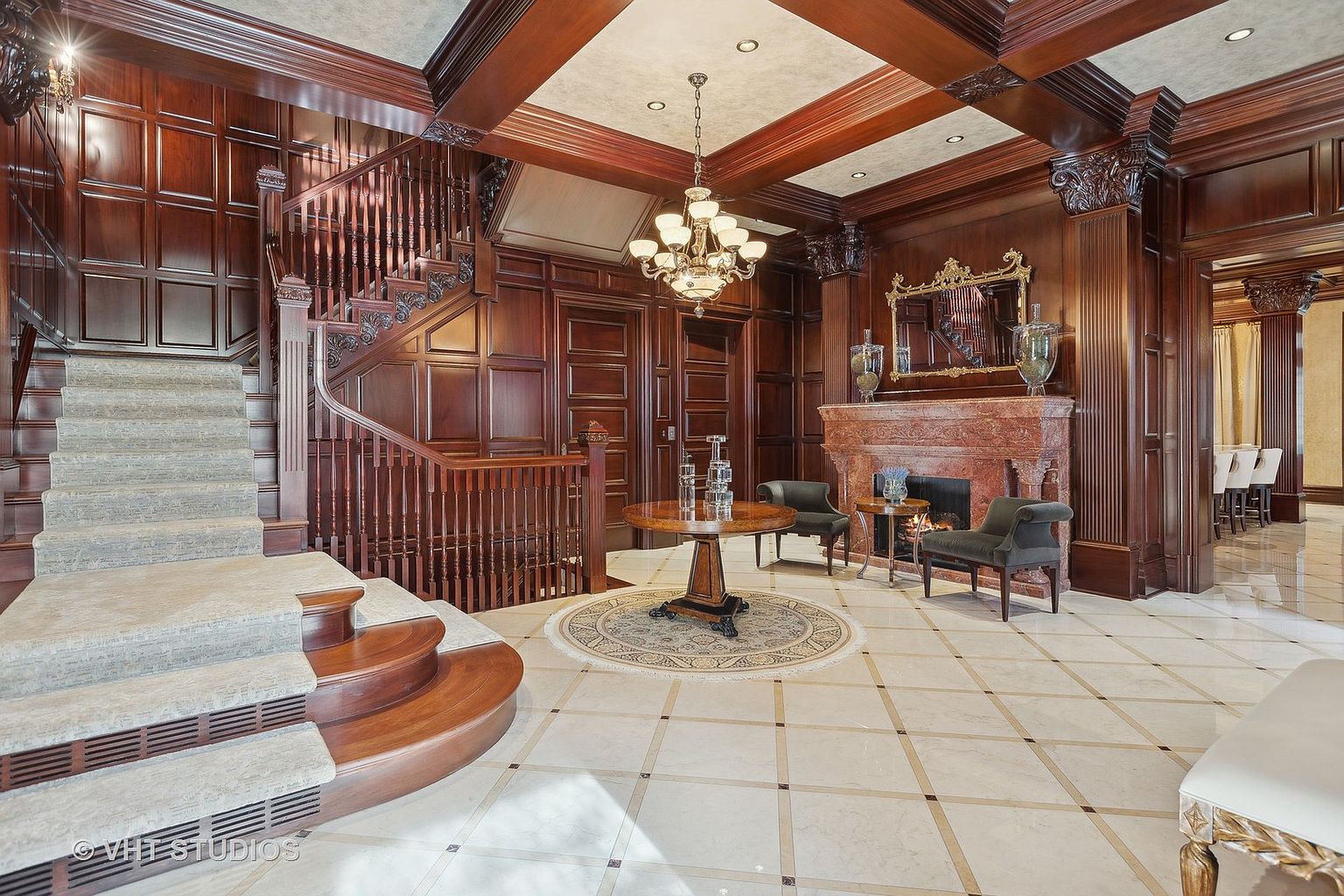 This grand foyer features rich wood paneling, a sweeping staircase with carpeted steps, and a decorative chandelier. A round rug sits beneath a central table, while a fireplace with ornate detailing adds warmth and character. The marble tiled floor and coffered ceiling contribute to the luxurious and elegant ambiance of the space.