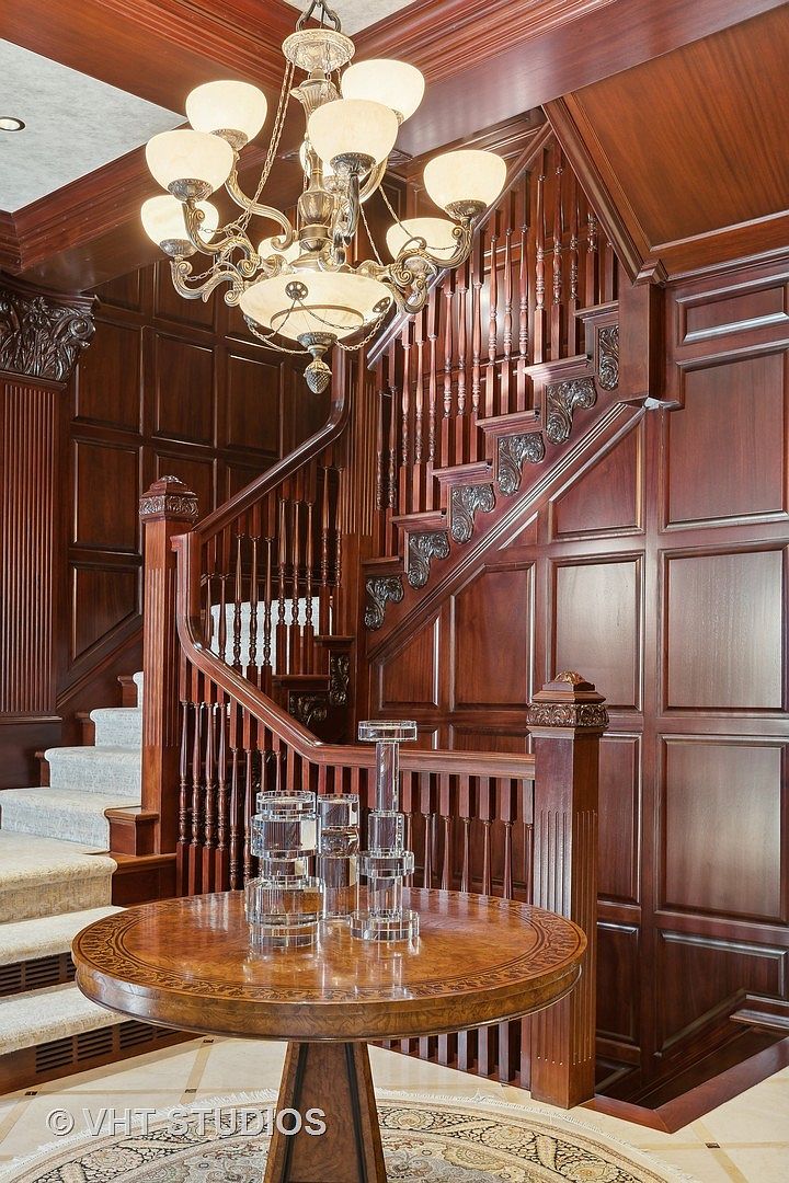 This interior shot showcases a grand hallway and staircase, featuring rich wood paneling and intricate detailing. A round table with decorative glass pieces sits in the foreground, while an ornate chandelier hangs above the staircase. The overall impression is one of luxury and classic elegance.