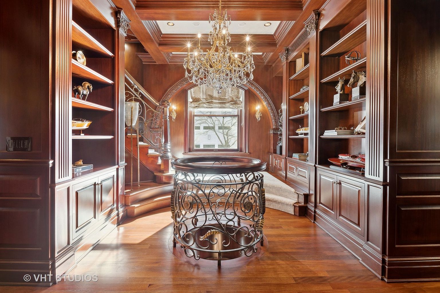 This is a luxurious home office or study featuring rich wood paneling and built-in bookshelves. A grand chandelier hangs from the coffered ceiling, illuminating the space. A decorative wrought iron table sits in the center of the room, and a staircase is visible in the background, adding to the room's grandeur.