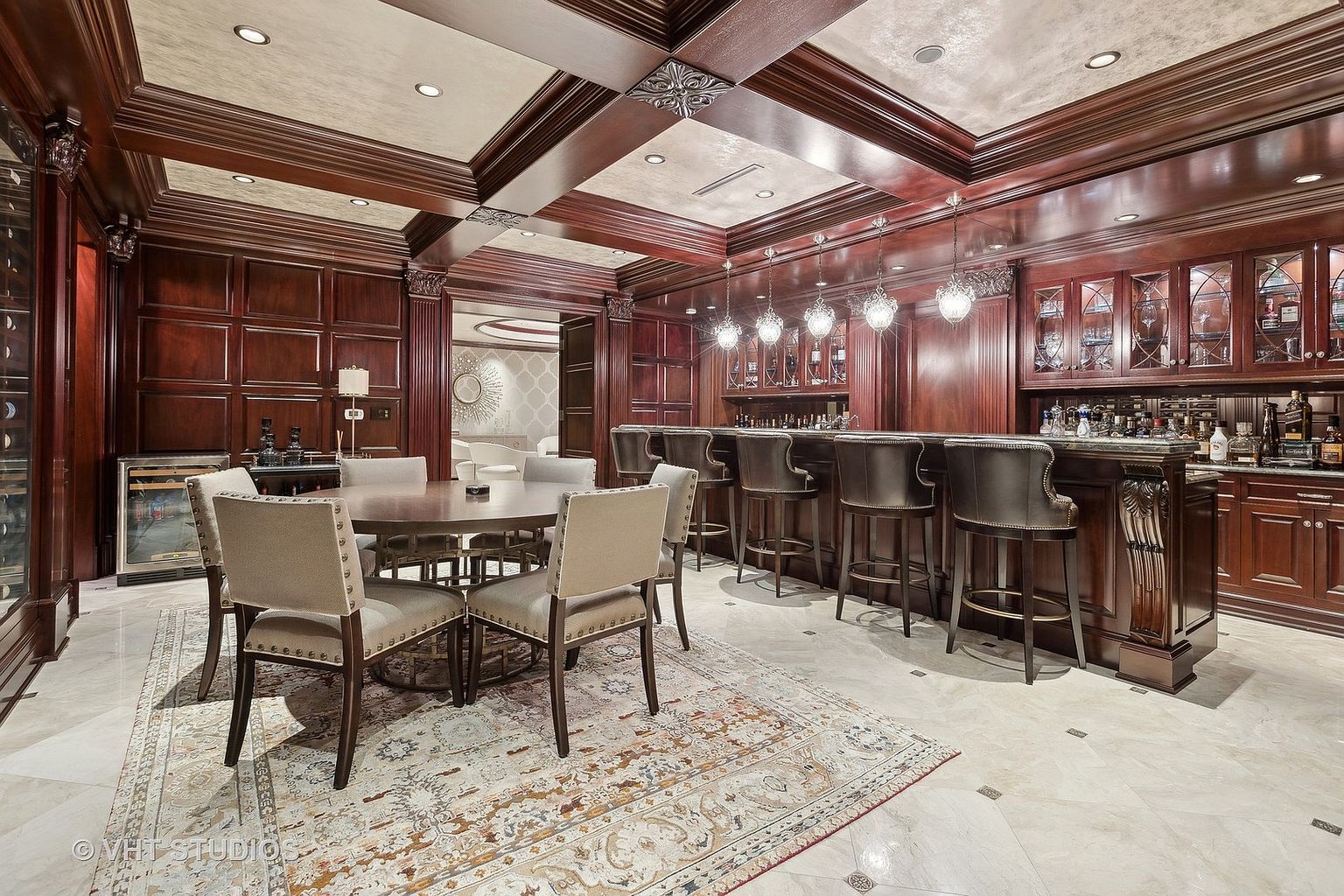 This is an interior shot of a luxurious wine cellar and bar area. The space features rich, dark wood paneling, cabinetry, and a coffered ceiling, creating a sophisticated and elegant atmosphere. A round dining table with upholstered chairs sits on an ornate rug, while a fully stocked bar with leather bar stools and pendant lighting provides a focal point for entertaining.