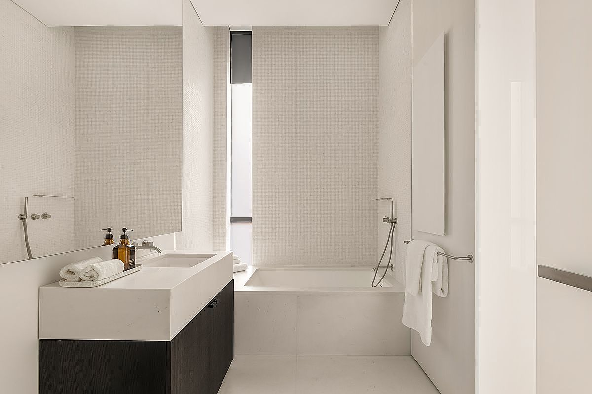 This minimalist bathroom features a sleek, wall-mounted vanity with a stone countertop and a dark wood base, paired with a large, frameless mirror. A deep soaking tub is positioned against a wall covered in small, light-toned mosaic tiles, complemented by a tall, narrow window that allows natural light to filter into the space. The overall aesthetic is clean, modern, and serene, emphasizing a neutral color palette and high-end finishes.