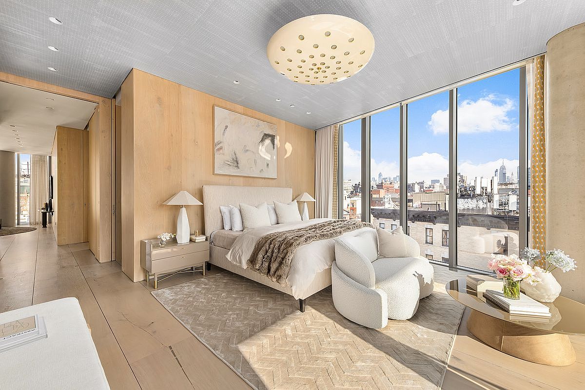 This sophisticated primary bedroom features light wood wall paneling and a textured ceiling, creating a warm and modern aesthetic. The room is anchored by a plush bed with neutral linens and a cozy throw, complemented by a curved armchair and a glass-topped coffee table. Large windows provide an expansive view of the city skyline, bathing the space in natural light.