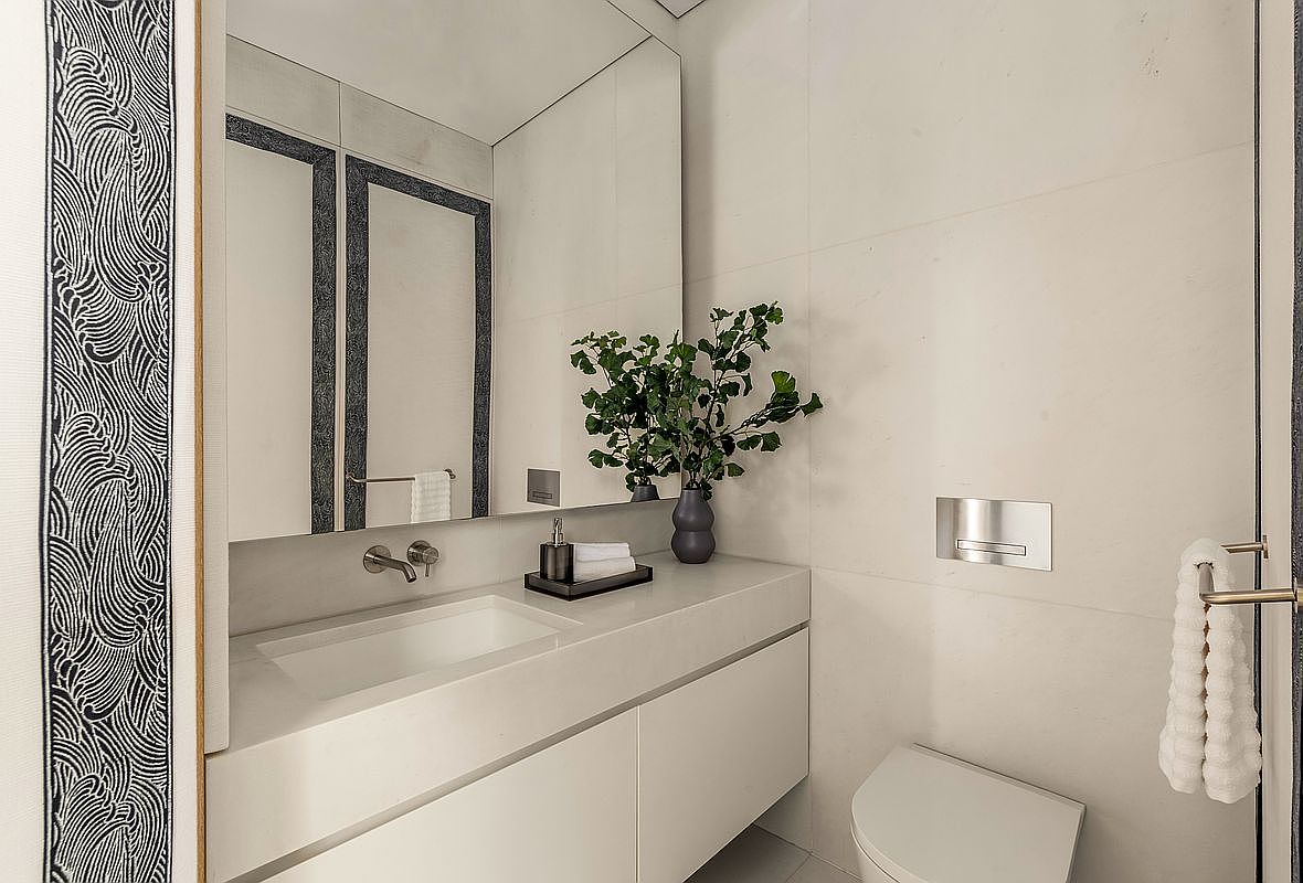 This modern powder room features a sleek, wall-mounted white vanity with a rectangular sink and minimalist faucet. A large, frameless mirror reflects the space, which is accented by a striking patterned wallpaper on the left wall and a decorative vase with greenery on the counter. The room is finished with light-toned wall tiles and a wall-mounted toilet, creating a clean and sophisticated aesthetic.