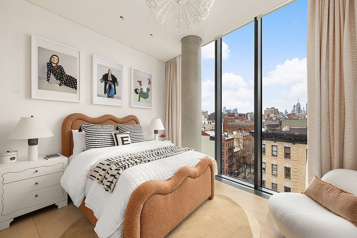 This modern bedroom features a textured, scalloped-edge bed frame in a warm earth tone, complemented by striped accent pillows and a neutral bedding ensemble. The room is bathed in natural light from expansive floor-to-ceiling windows that offer a panoramic view of the city skyline. A white nightstand with a contemporary lamp and a circular area rug complete the clean, sophisticated aesthetic.