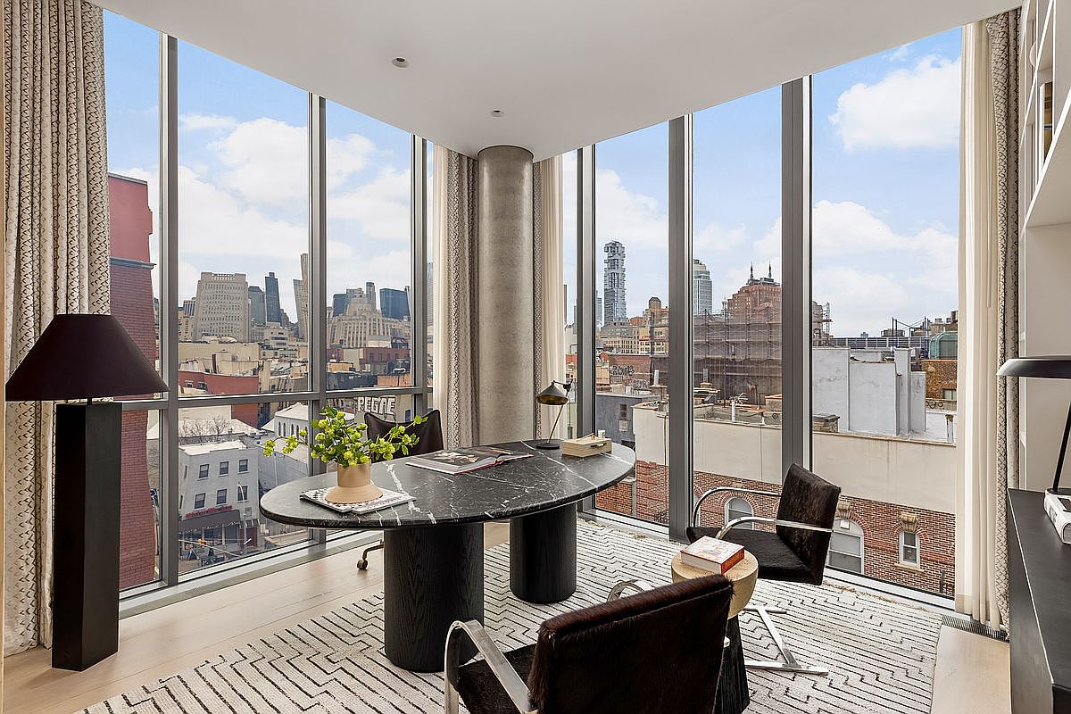 This sophisticated home office features a striking oval-shaped desk with a dark stone top and cylindrical bases, positioned centrally to take advantage of the expansive city views. The room is framed by floor-to-ceiling windows that provide abundant natural light and a panoramic perspective of the urban skyline. A patterned area rug anchors the space, which is furnished with modern chairs and a tall, minimalist floor lamp, creating a professional yet elegant atmosphere.