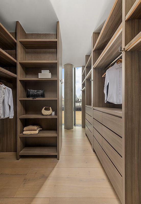 This modern walk-in closet features custom wood-toned shelving and cabinetry, providing ample storage space for clothing and accessories. The layout includes open shelving on the left and a combination of hanging rods and drawers on the right, all set against a neutral, minimalist backdrop. A central pillar leads the eye toward a large window at the end of the hallway, which offers a view of the city skyline.